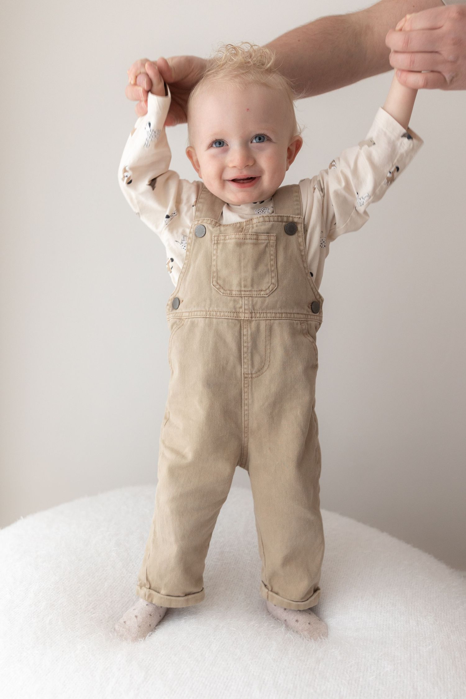 Baby in beige overalls standing on white surface with supporting hands.
