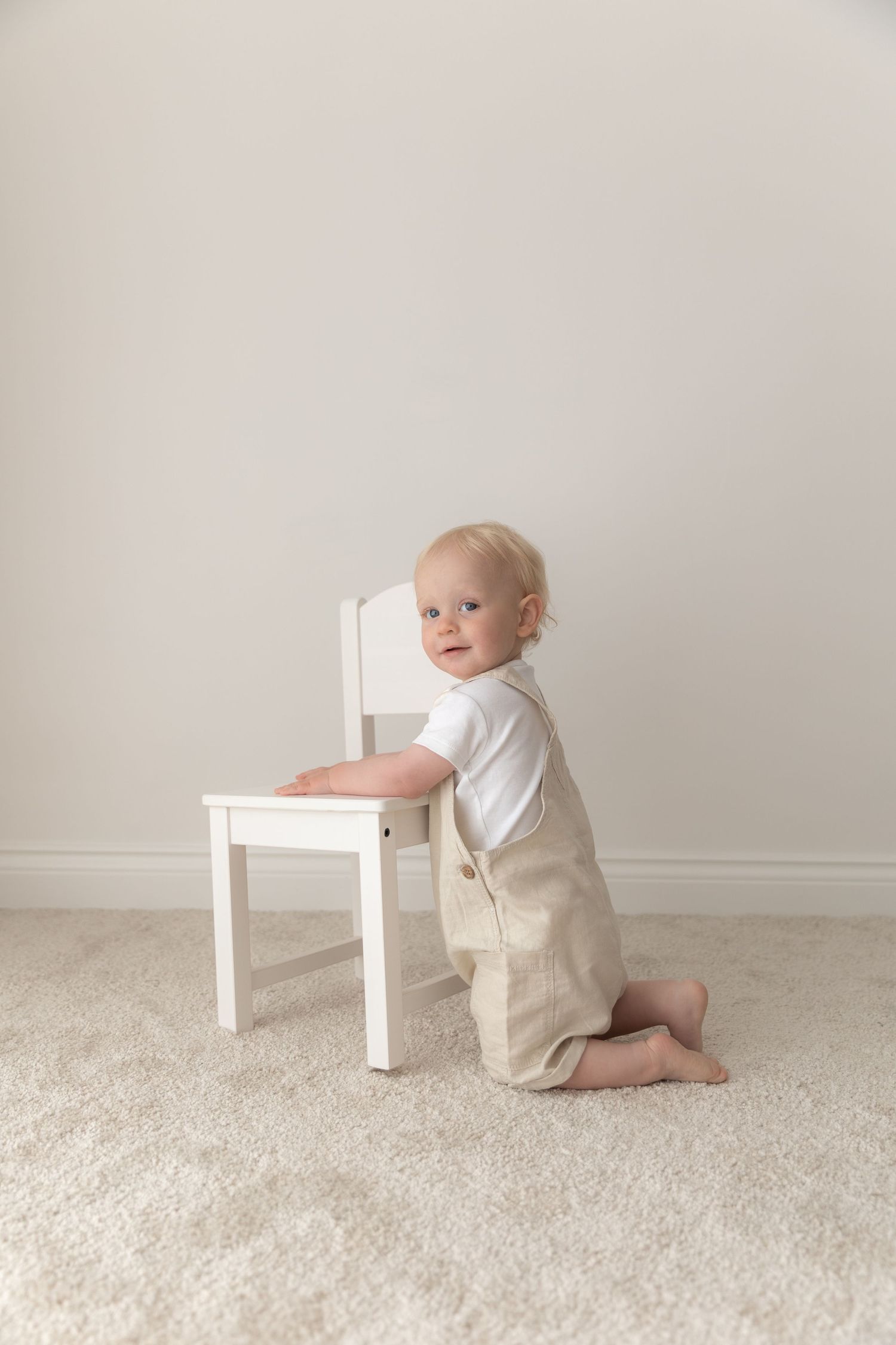 A small toddler in tan overalls and white shirt kneels next to a white wooden chair on beige carpet.