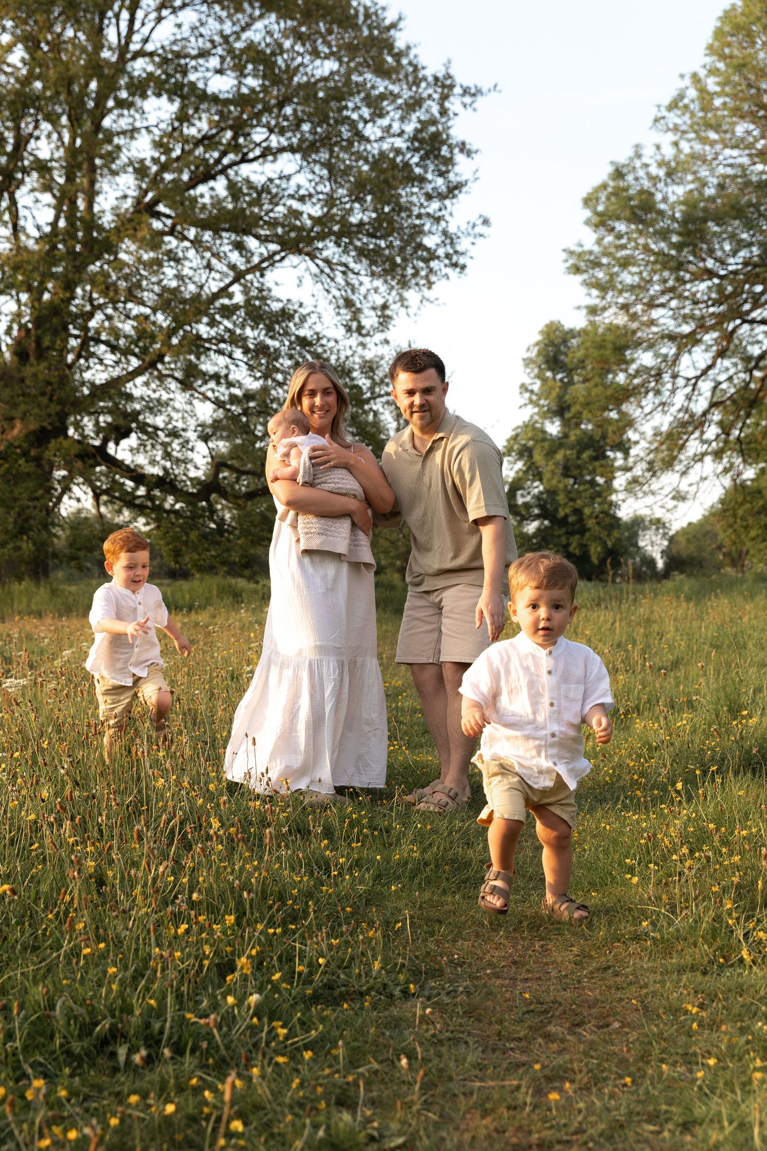 A family stands together in a sunny meadow with wildflowers during golden hour, wearing light summer clothing.