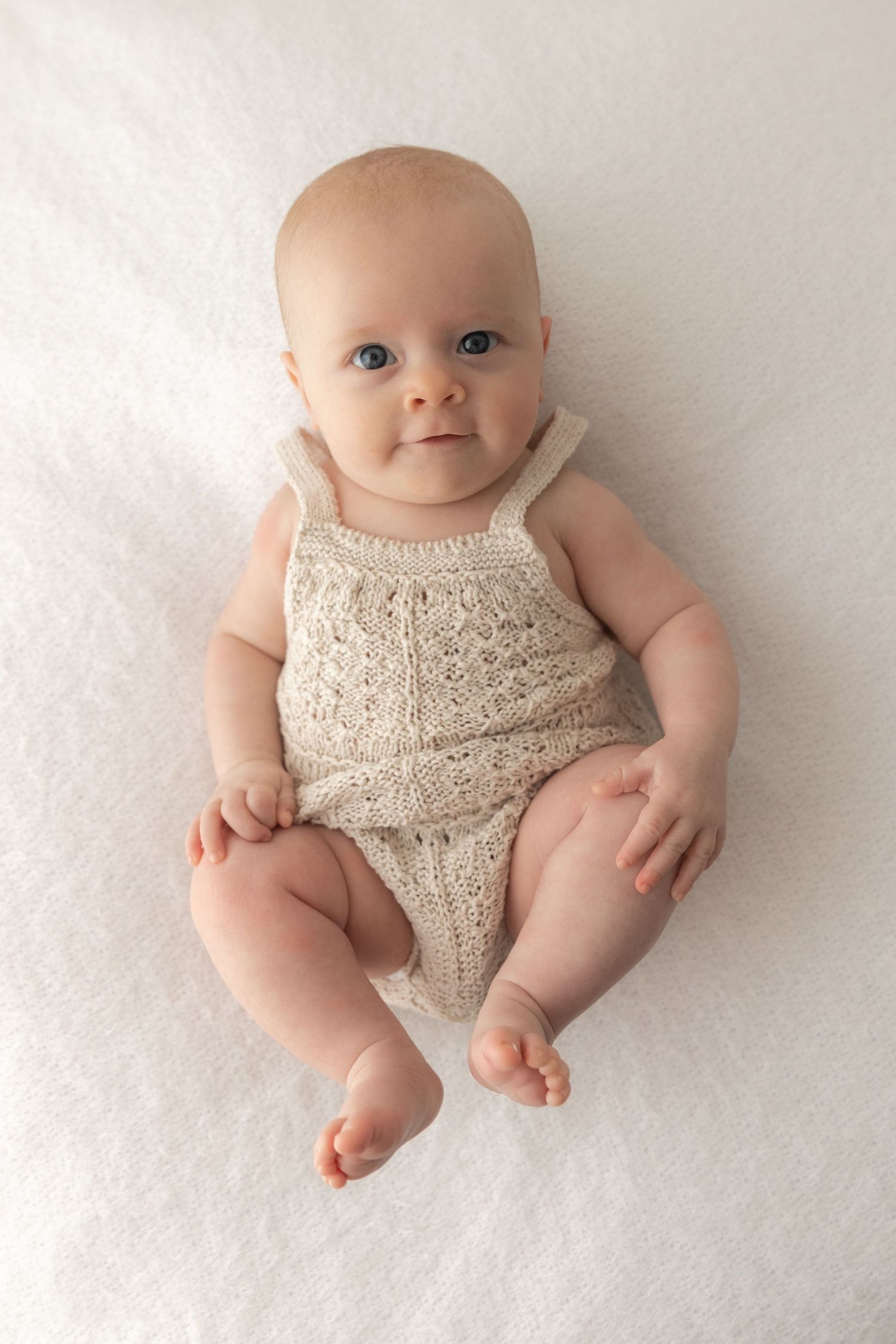 Baby in cream knit romper sitting on white background with bright eyes.
