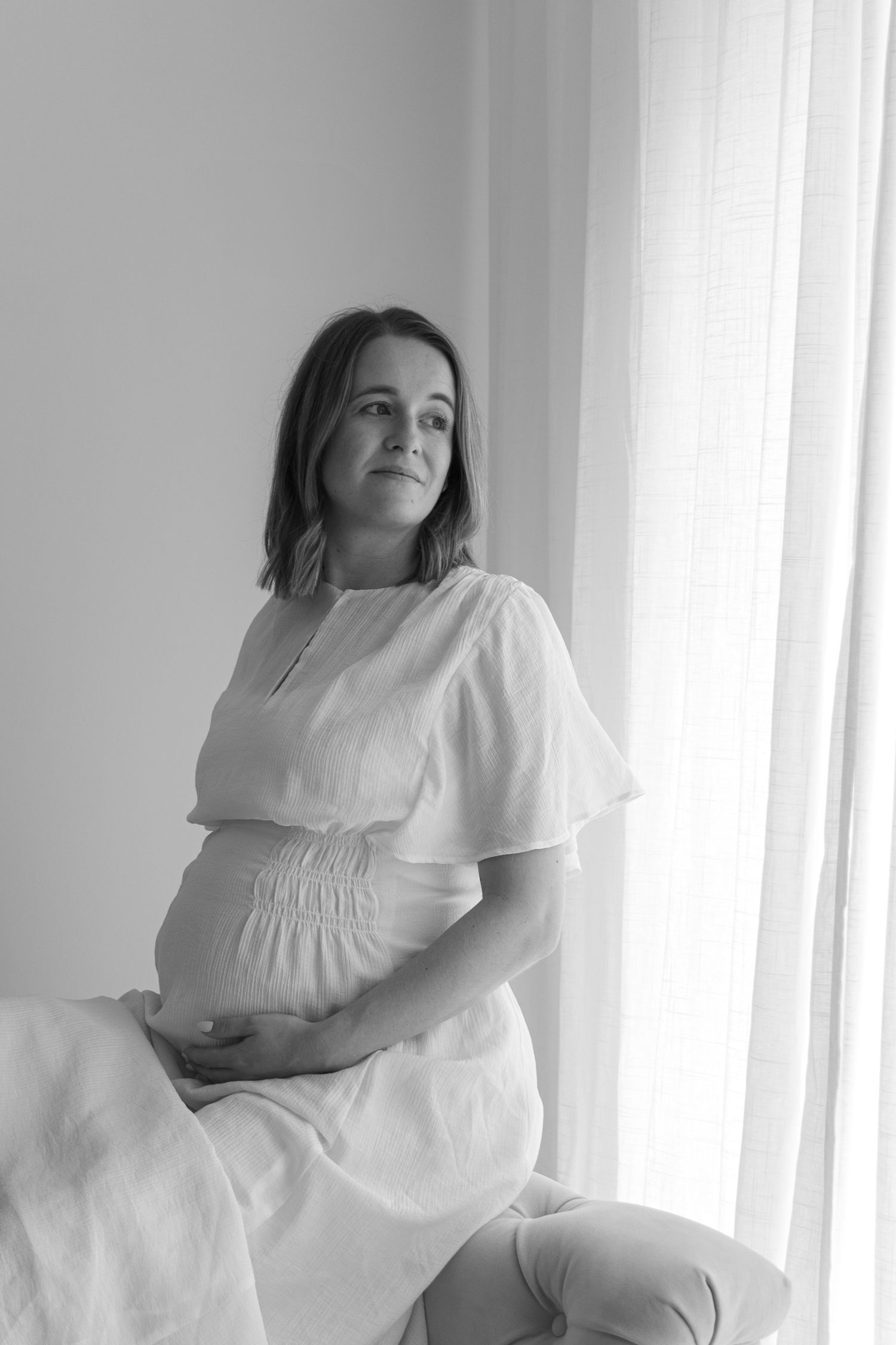 Black and white maternity portrait of woman in white dress seated by window.