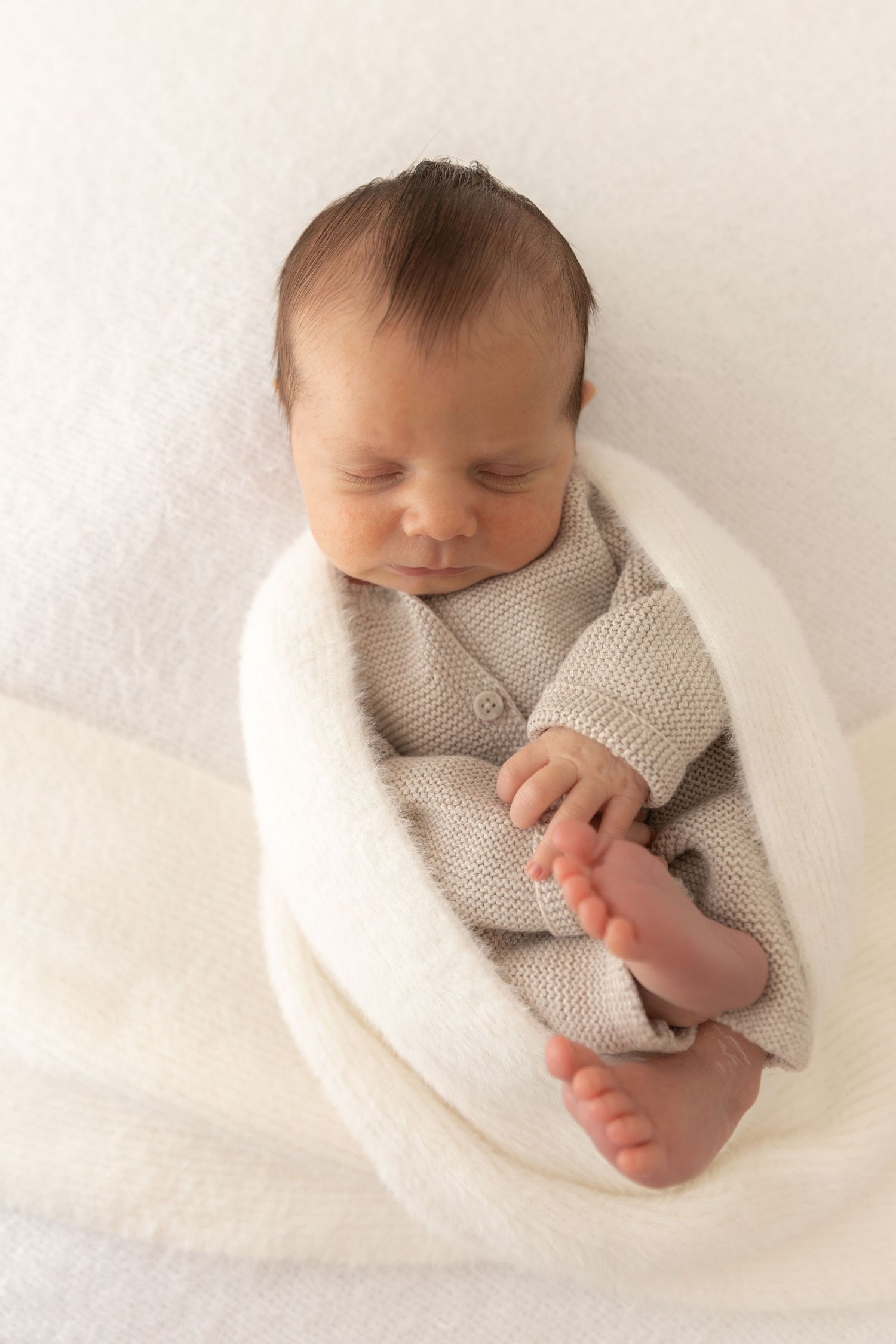 Sleeping newborn wearing cozy gray knit sweater swaddled in soft white blanket during professional photo session.