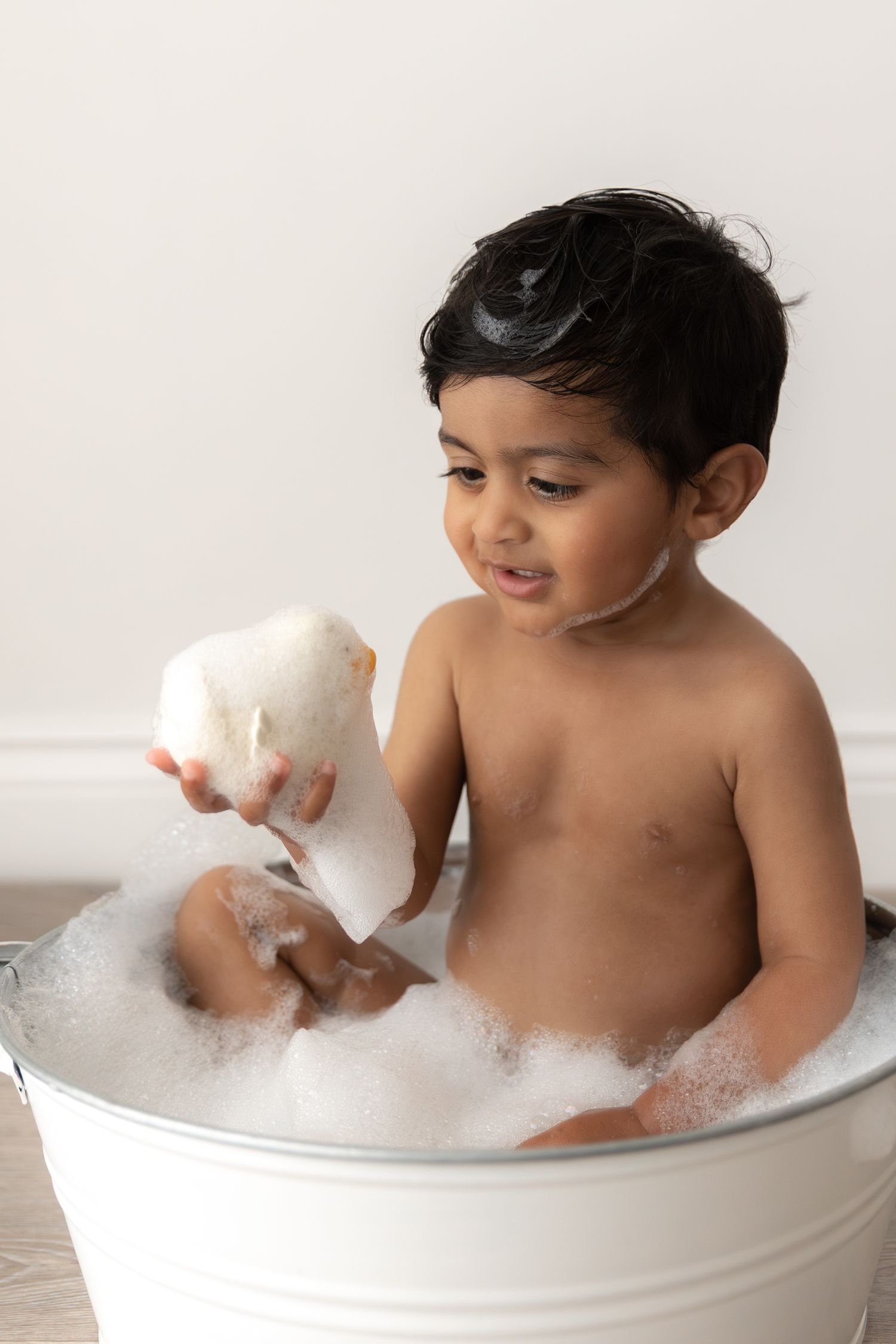 Young child enjoying bathtime in a white bucket tub while playing with a white sponge toy.