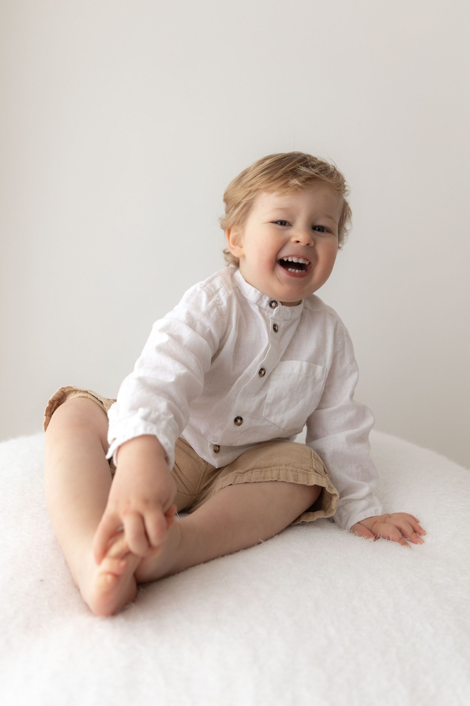 Joyful toddler in white shirt and khaki shorts sitting on white surface.