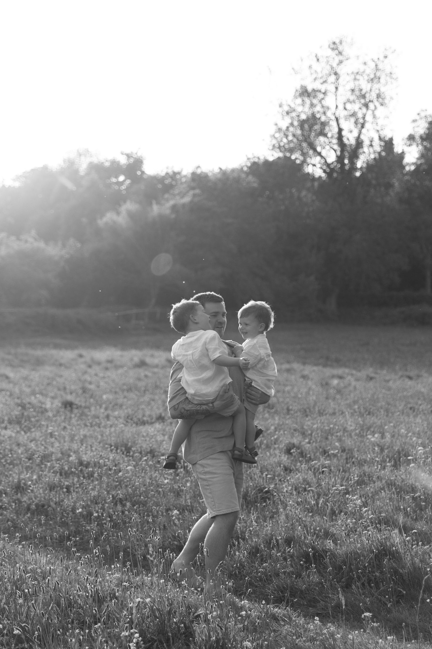 Black and white photo of a parent holding two children in a sunlit field at sunset.