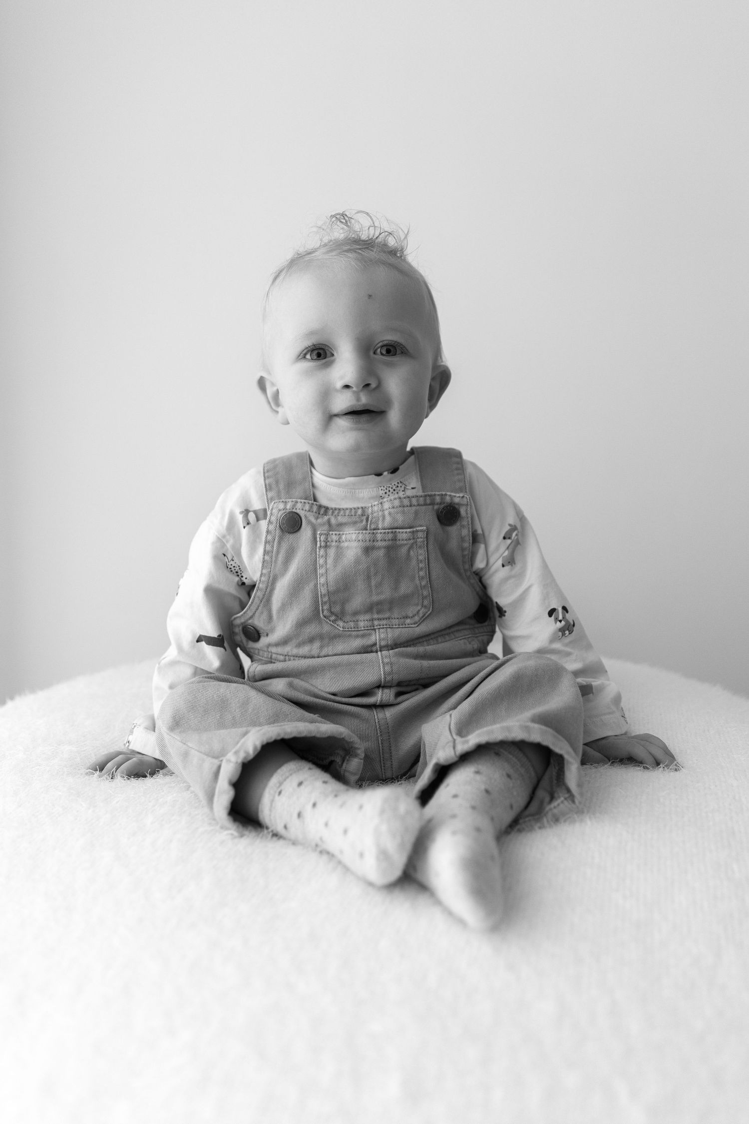 Baby in denim overalls sitting on white surface against neutral background.
