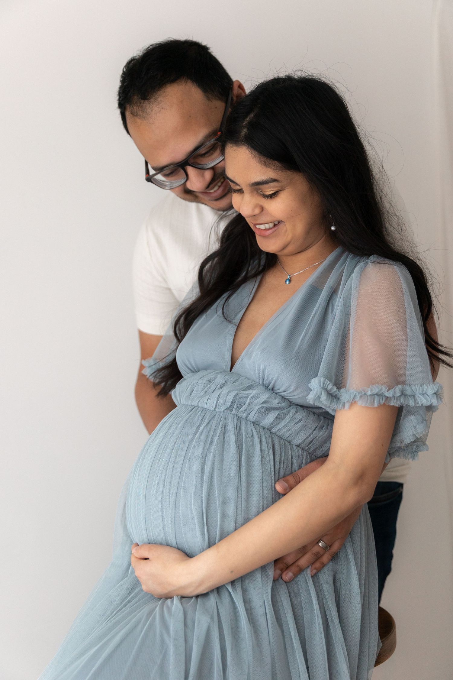 Expectant couple in intimate maternity photoshoot wearing light blue dress and white shirt.