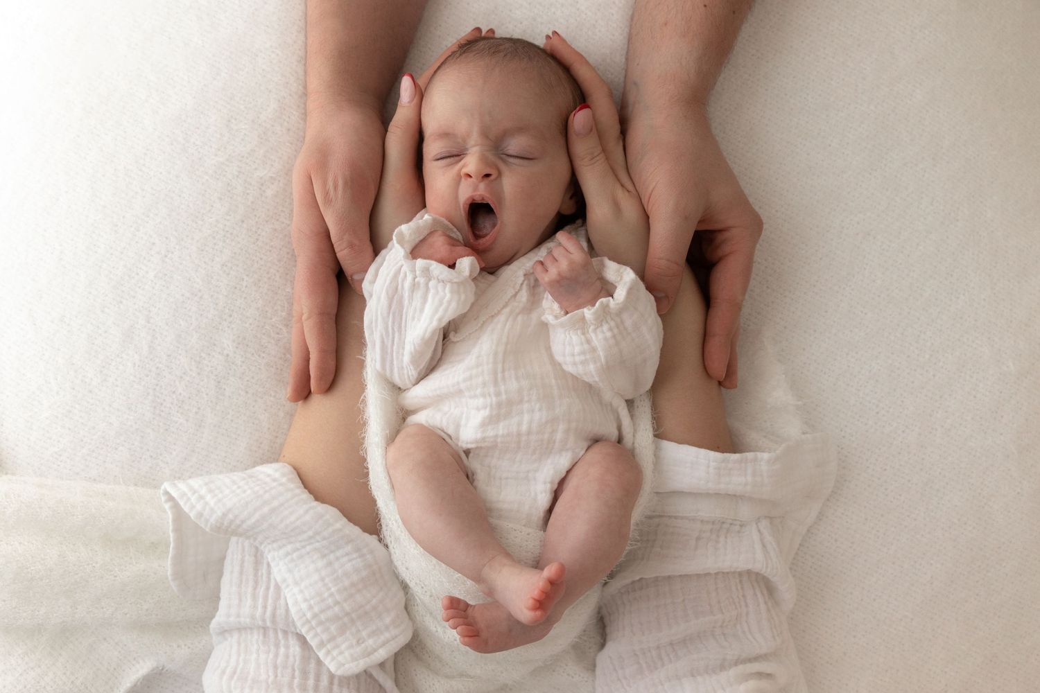 Newborn baby in white outfit yawning while being held on white textured blanket.
