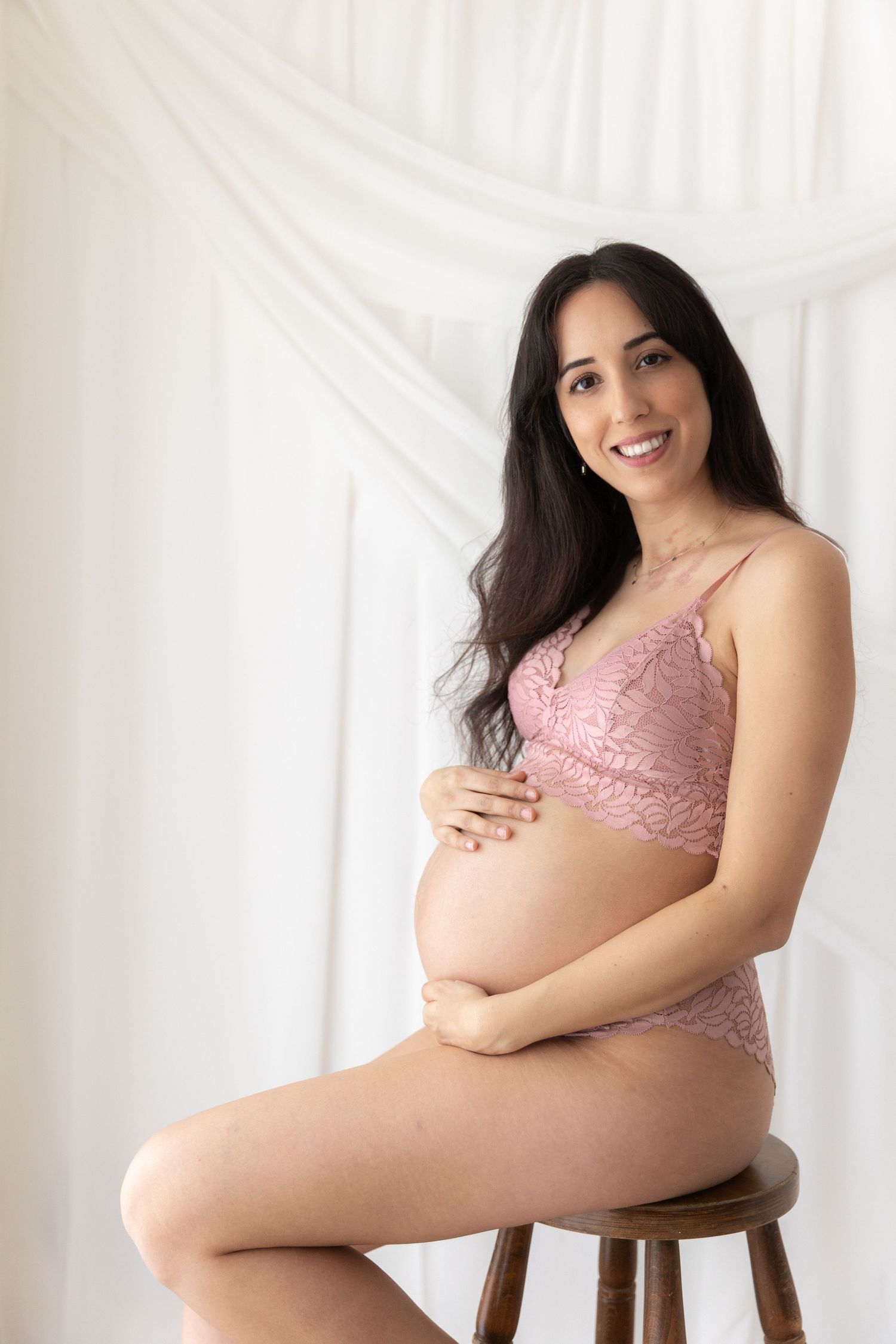Maternity photo of woman in pink lingerie sitting on wooden stool.