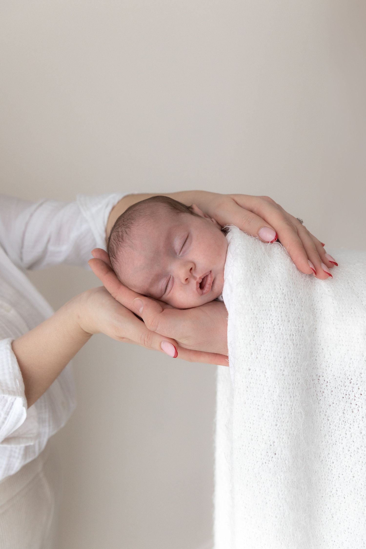 Sleeping newborn baby cradled in gentle hands against soft white fabric background.