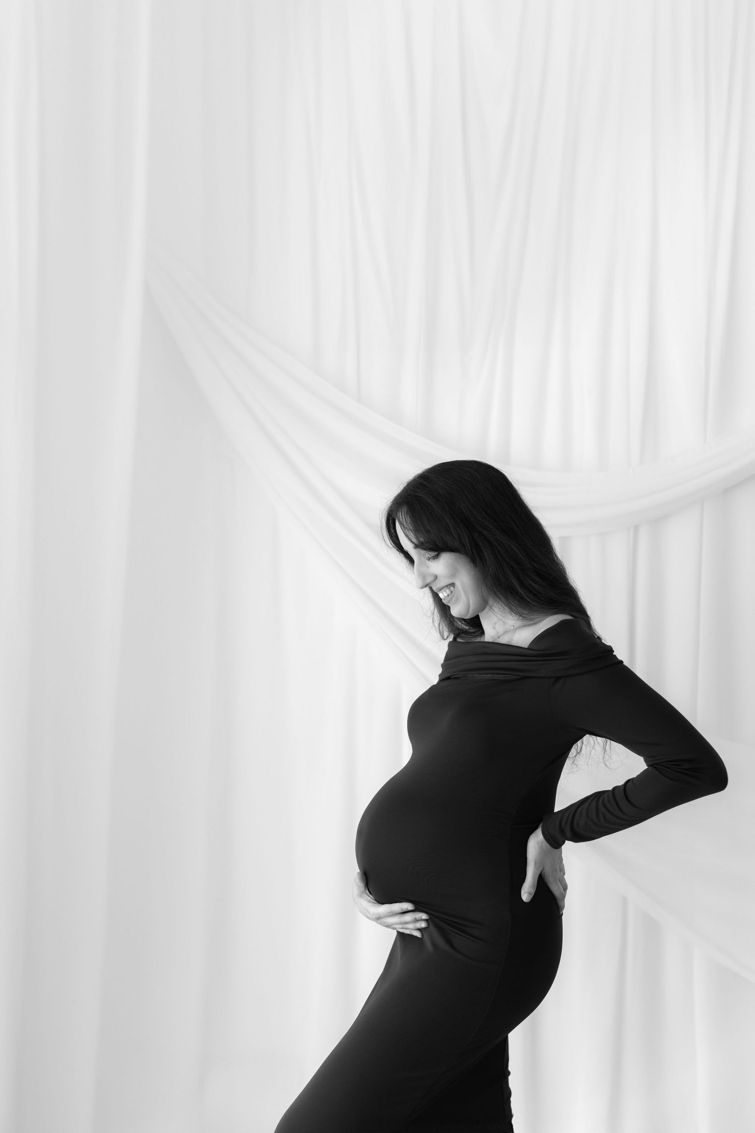 Artistic black and white silhouette of pregnant woman in form-fitting dress against curtains.