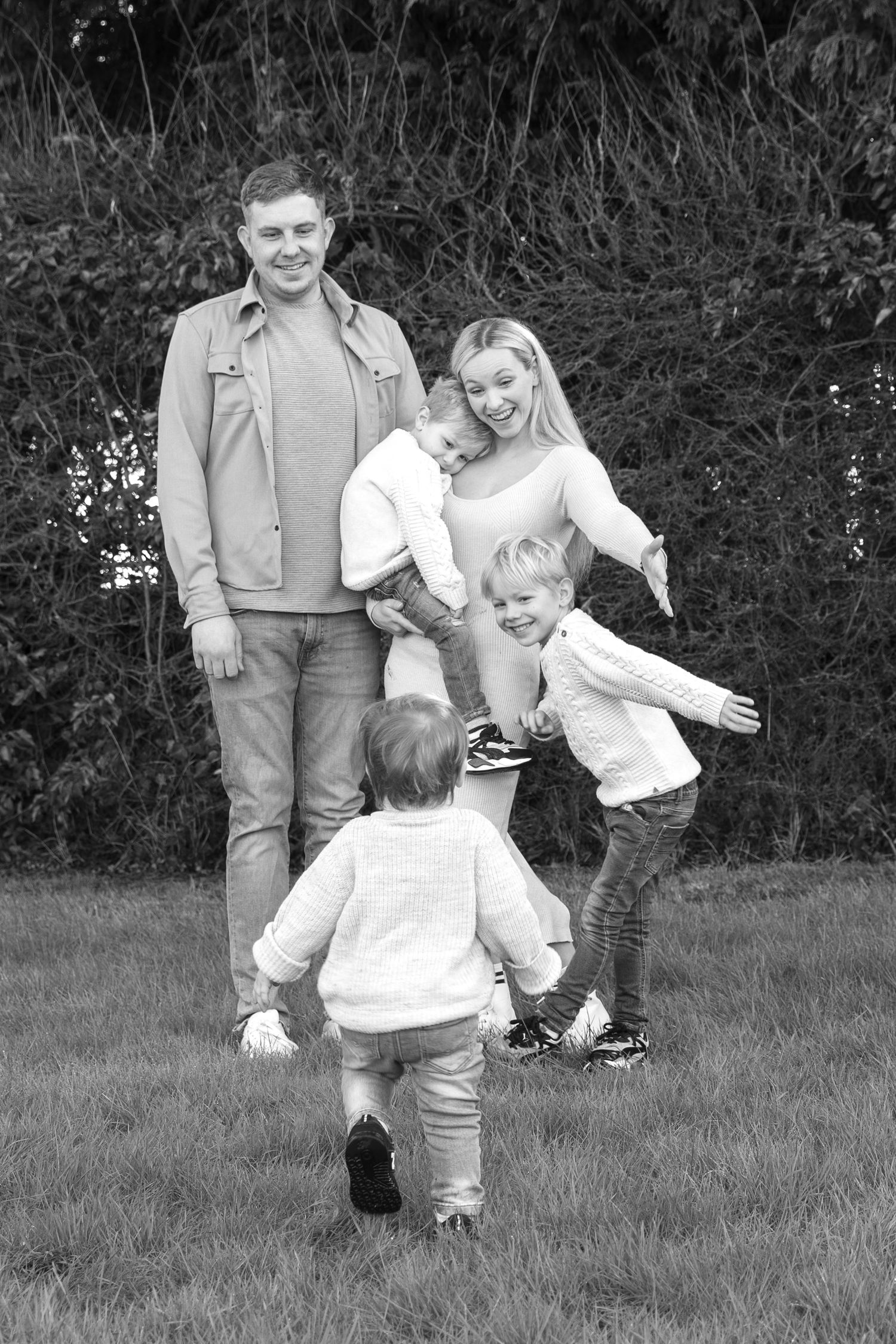 Black and white family portrait of parents and children playing together outdoors in front of bushes.