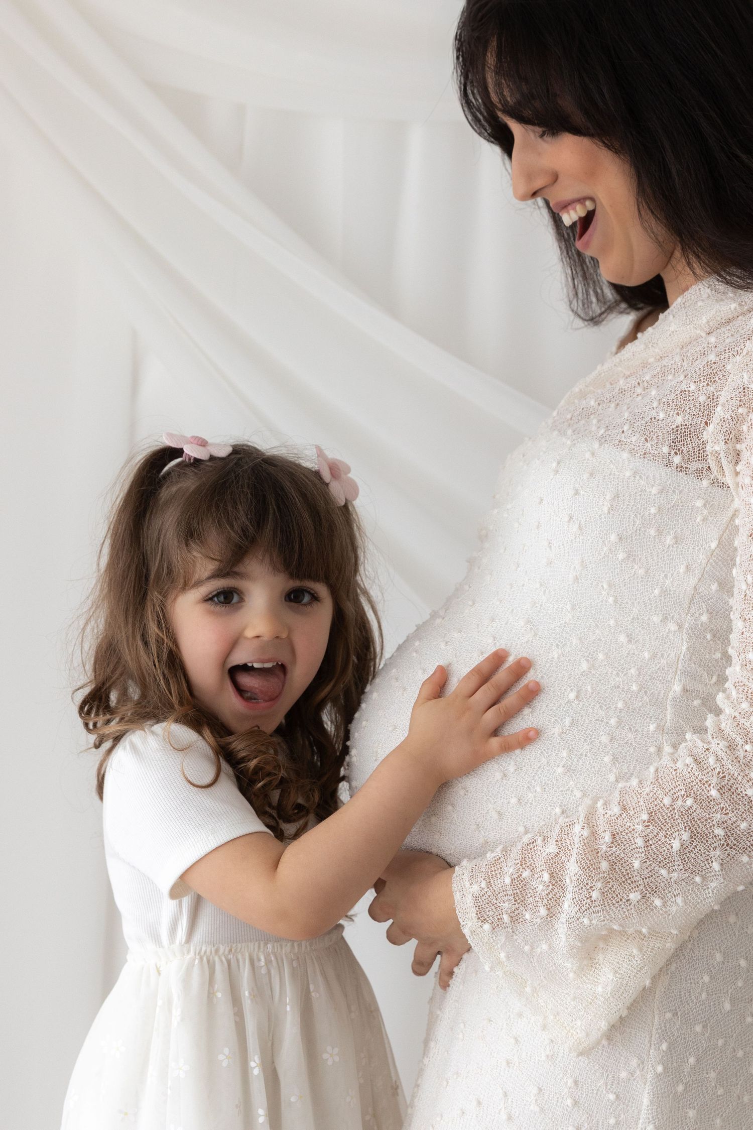 Joyful moment between mother and excited child touching a pregnant belly in white lace dress.