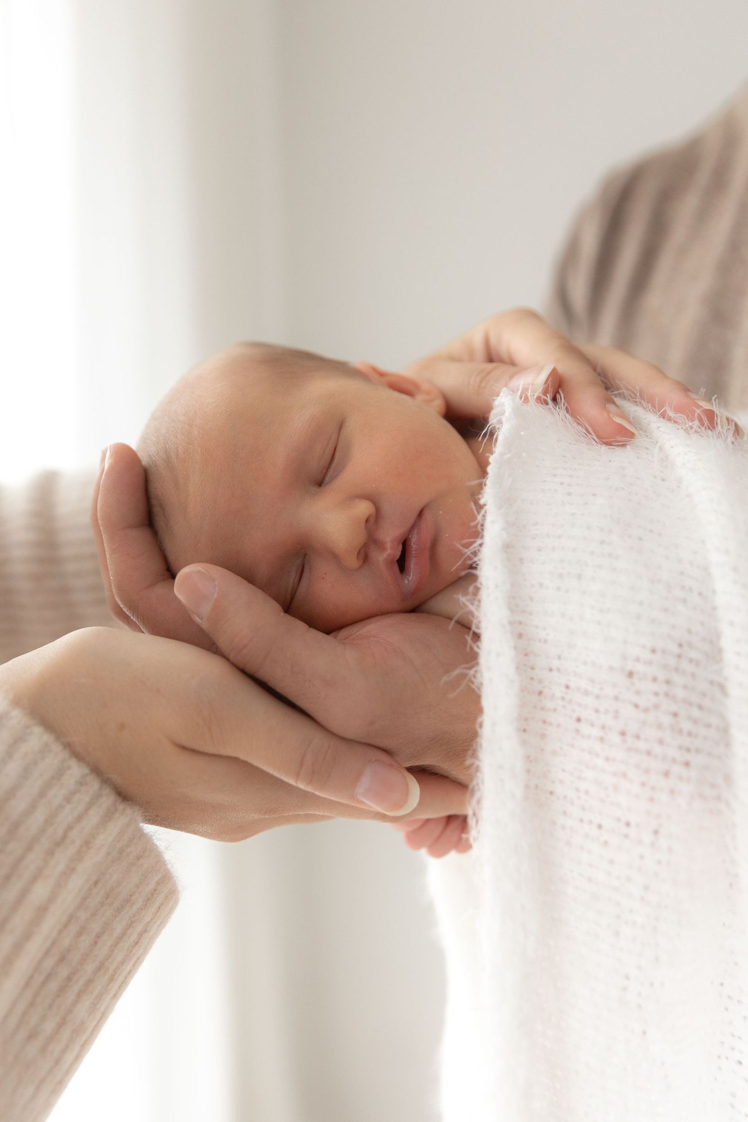 Delicate newborn sleeping peacefully in gentle hands wrapped in soft white fabric.
