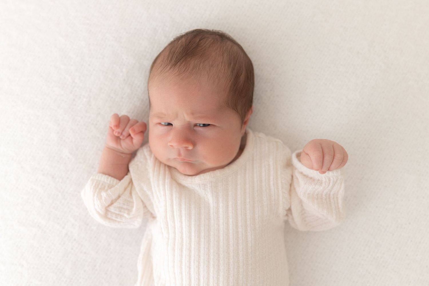 Baby wearing white ribbed sweater lying on light background with arms raised.