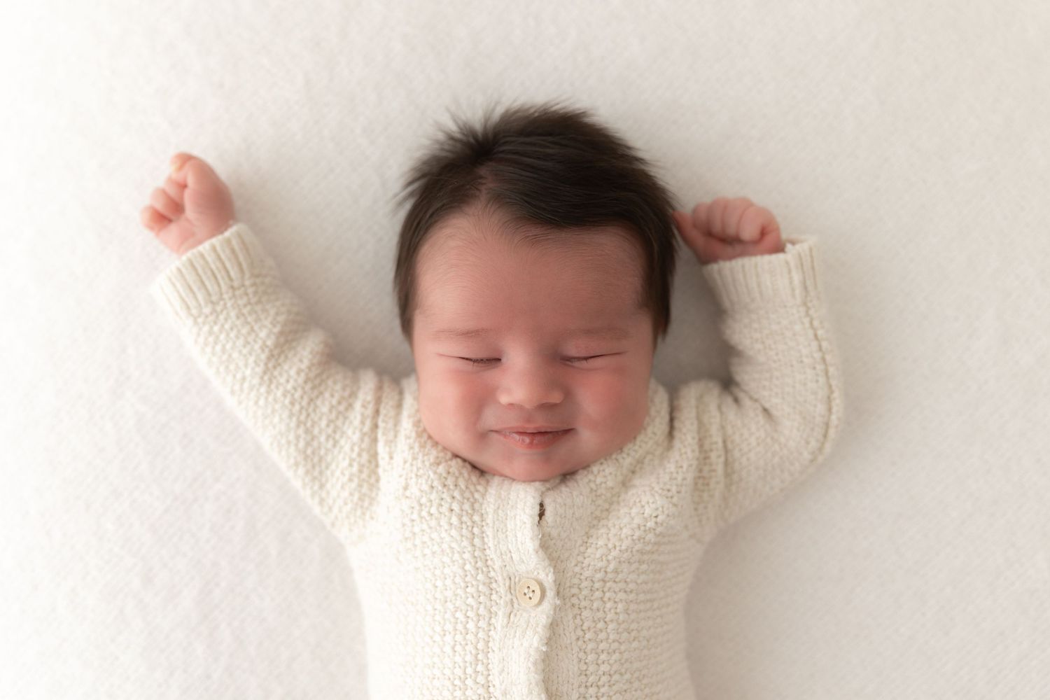 Smiling newborn baby wearing white knitted cardigan stretches arms on white background.