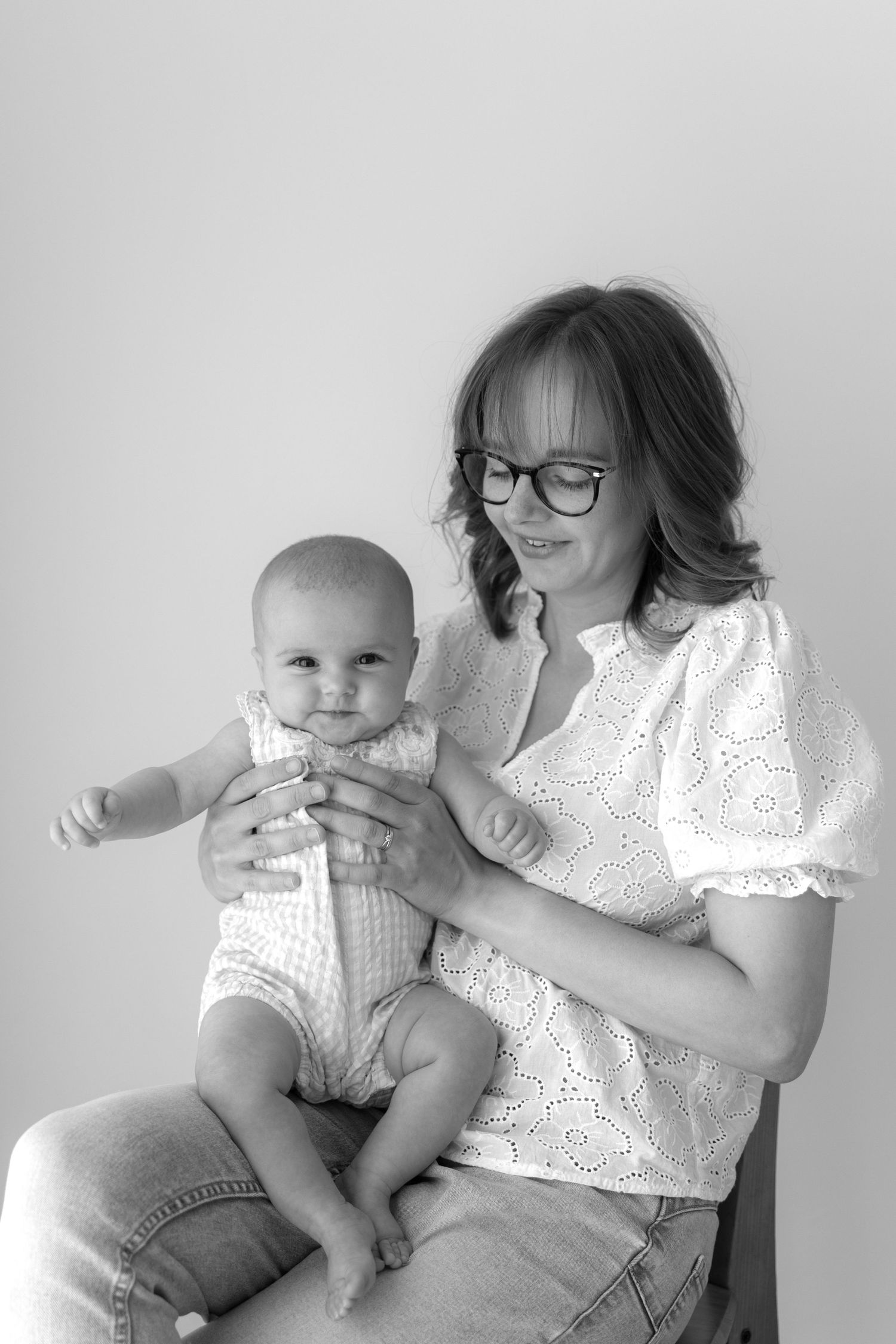 Mother in eyeglasses holding baby in white outfit against grey background.