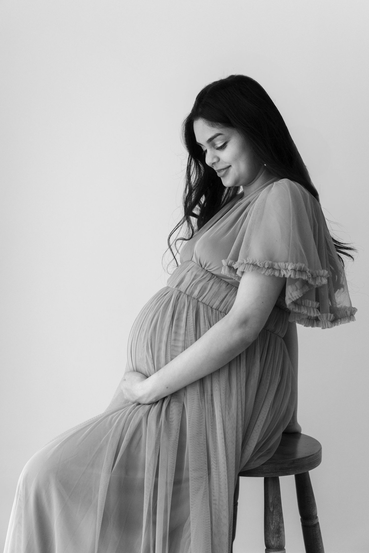 Black and white portrait of an expectant mother in a flowing dress sitting on a stool.