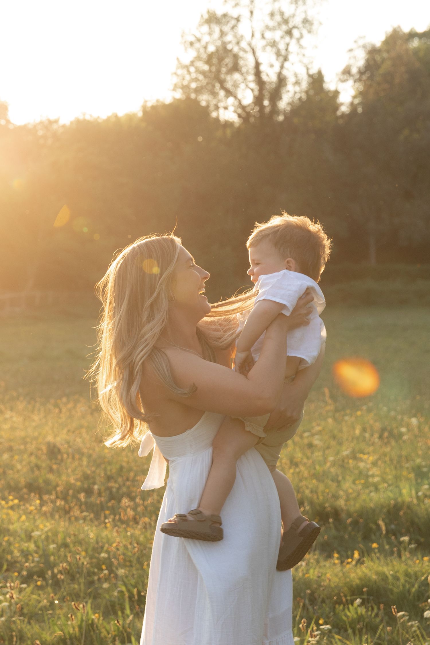 A tender moment captured during golden hour in a grassy field with sunlit silhouettes against a blurred forest backdrop.