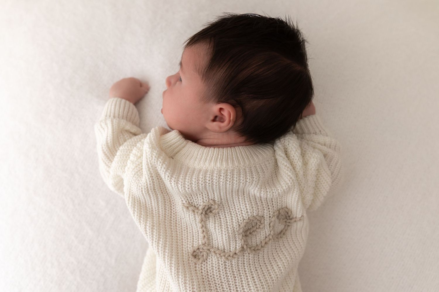 Infant wearing cozy white knit sweater resting on soft white surface in gentle lighting.