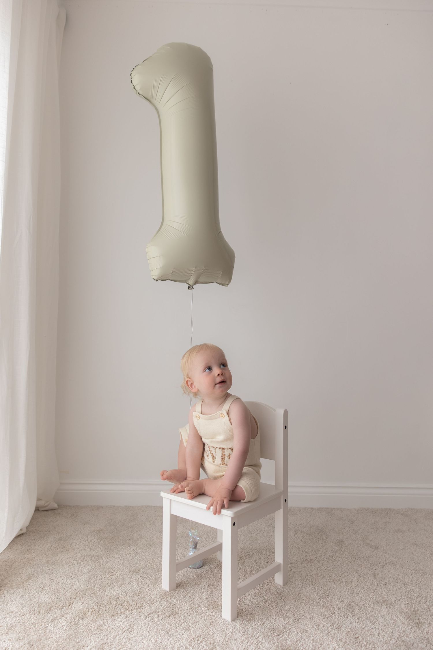 A baby in white sits on a small chair looking up at a large number one balloon floating above.