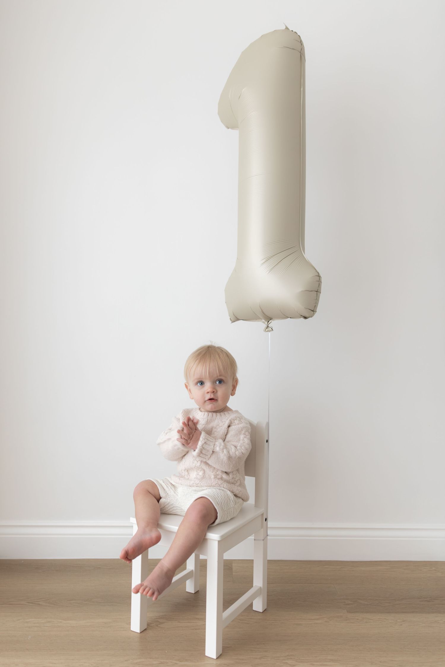 Baby sitting on white chair beneath large number one balloon for first birthday photo.