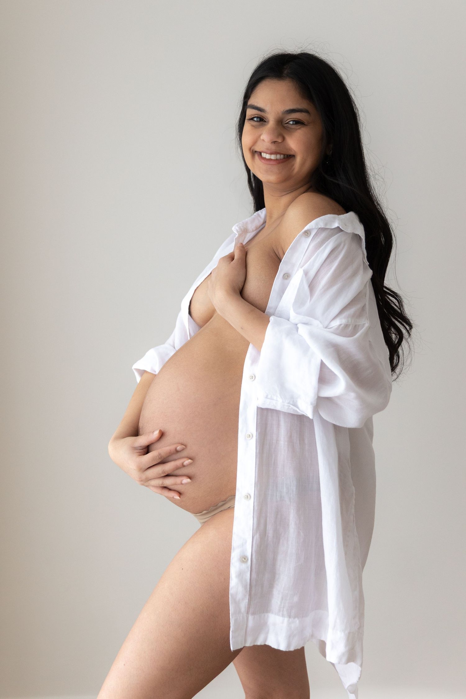 Side view of pregnant woman in white button down shirt posing against light backdrop.