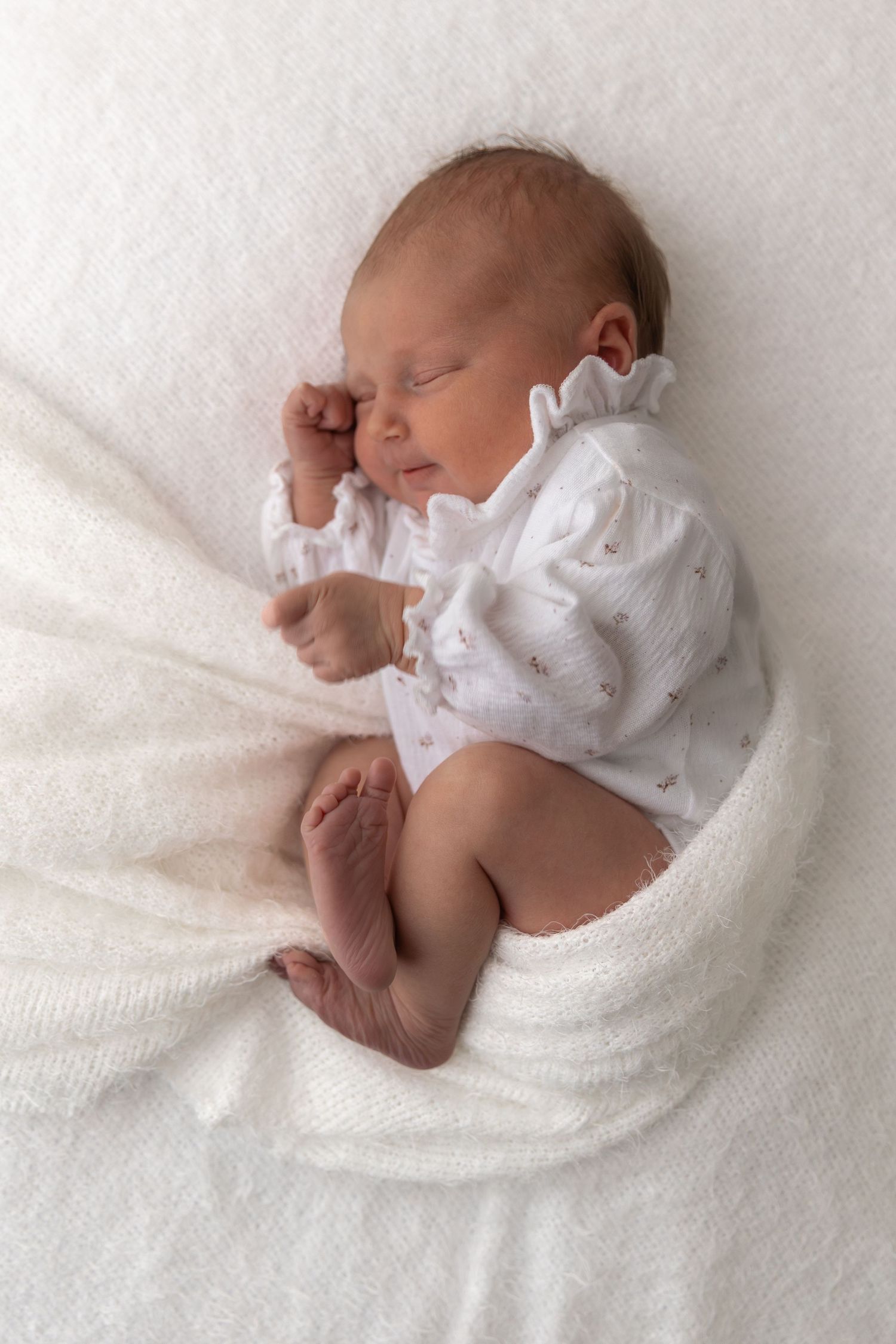 Sleeping newborn in white ruffled outfit rests peacefully on soft white blanket.
