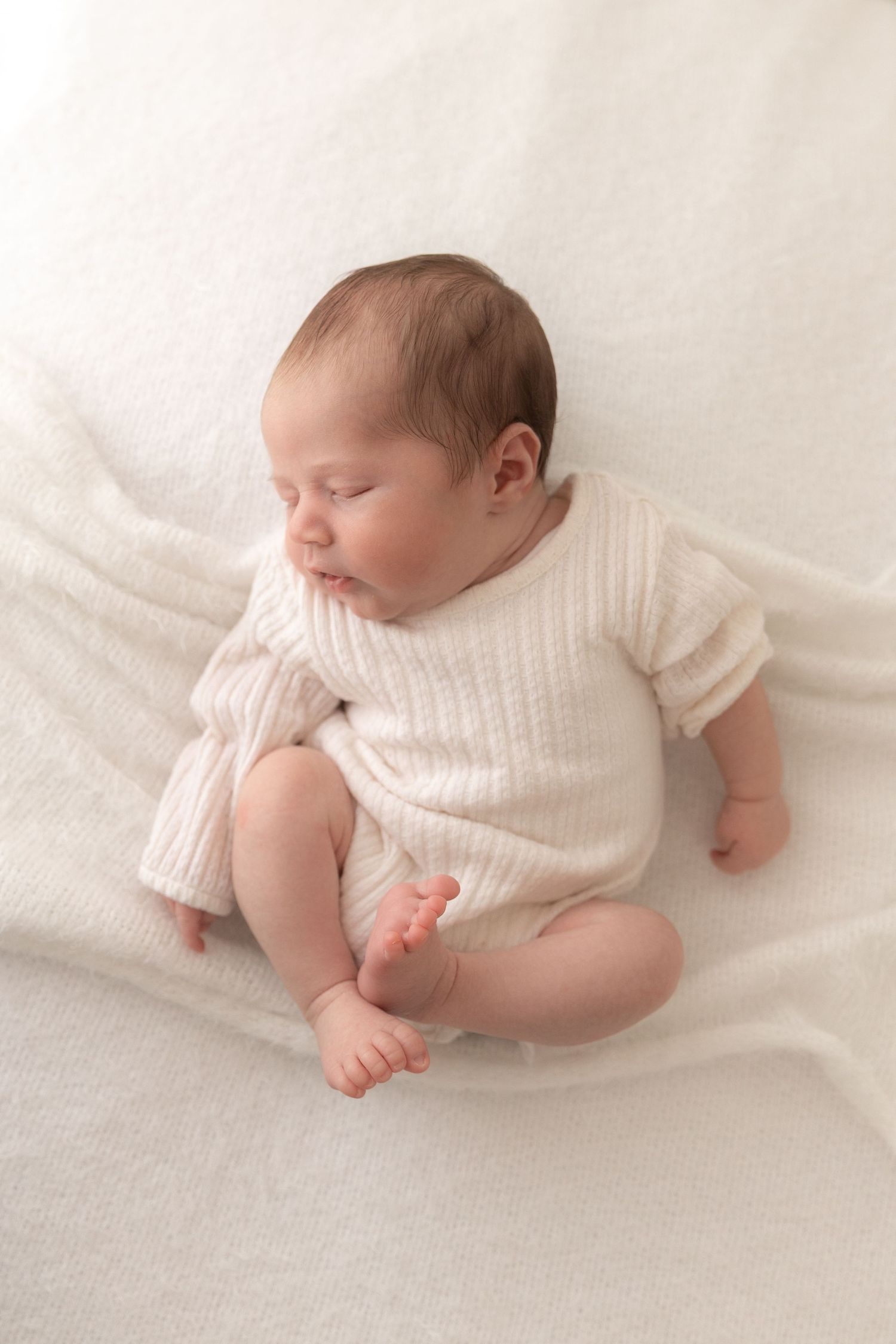 A peaceful sleeping baby wearing a soft white sweater lies on cream-colored bedding.