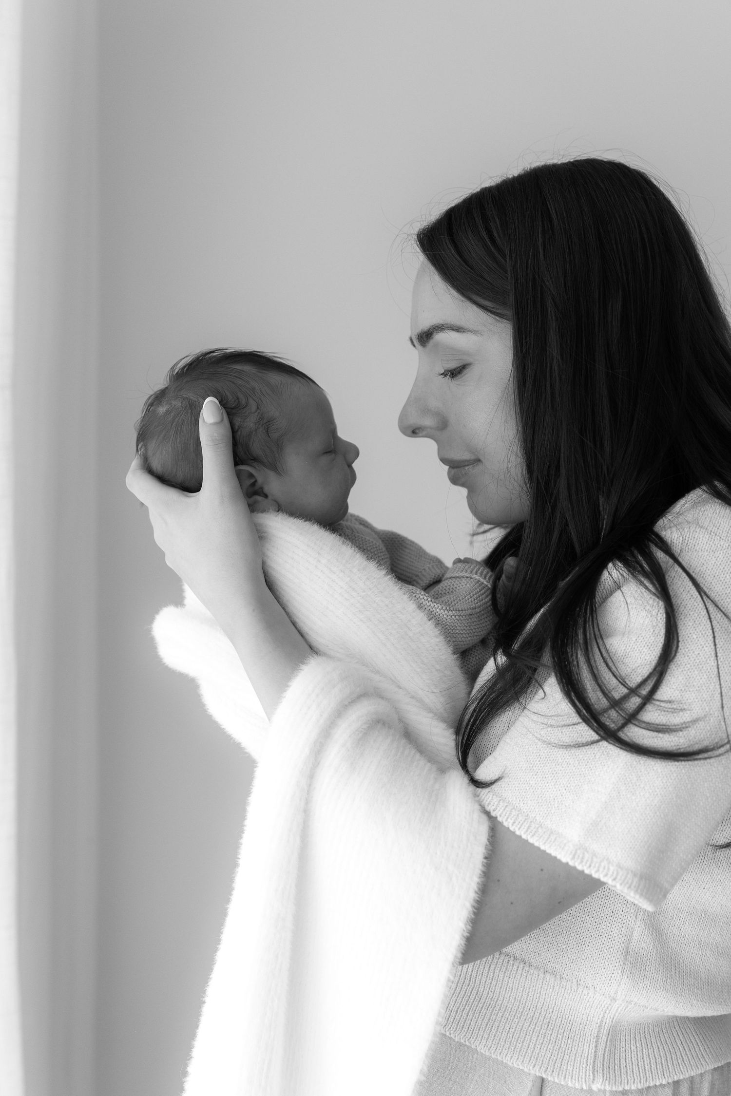 Woman in white sweater holding swaddled newborn baby while looking down with tender expression in black and white portrait.