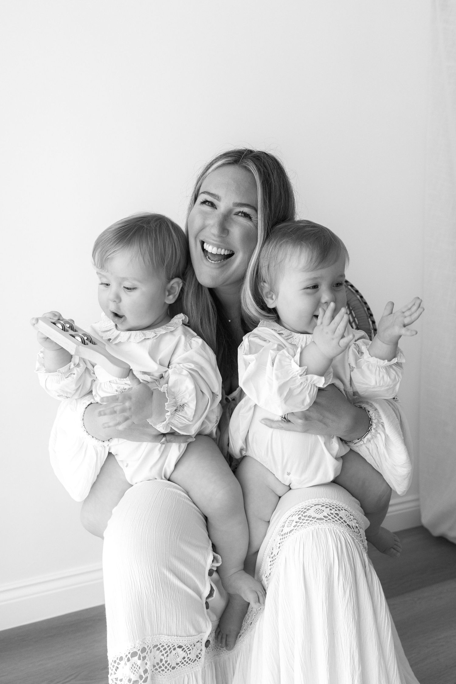 Mother holding twin babies in white dresses against neutral wall.