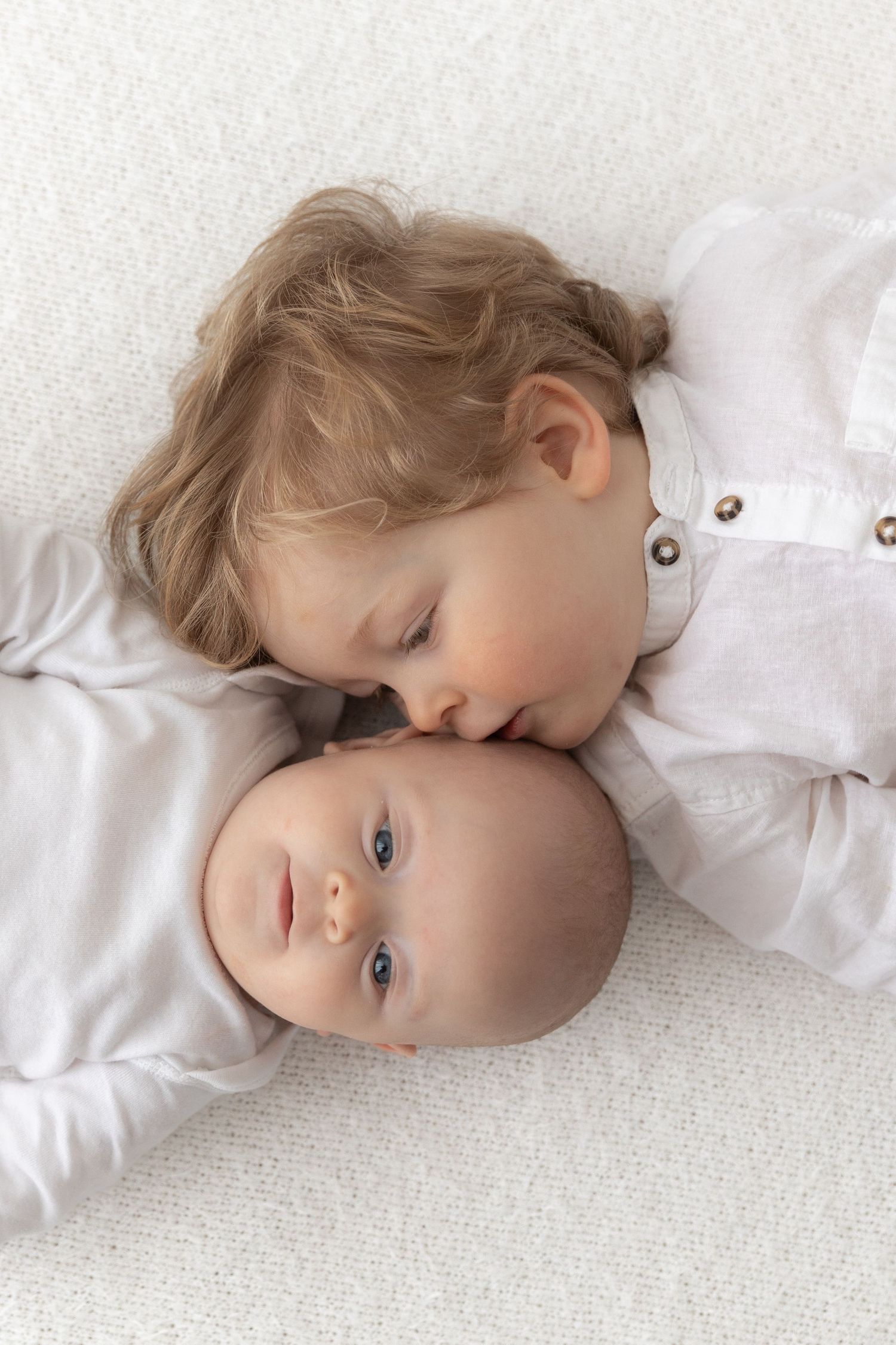 Two siblings lying close together on white fabric in gentle portrait.