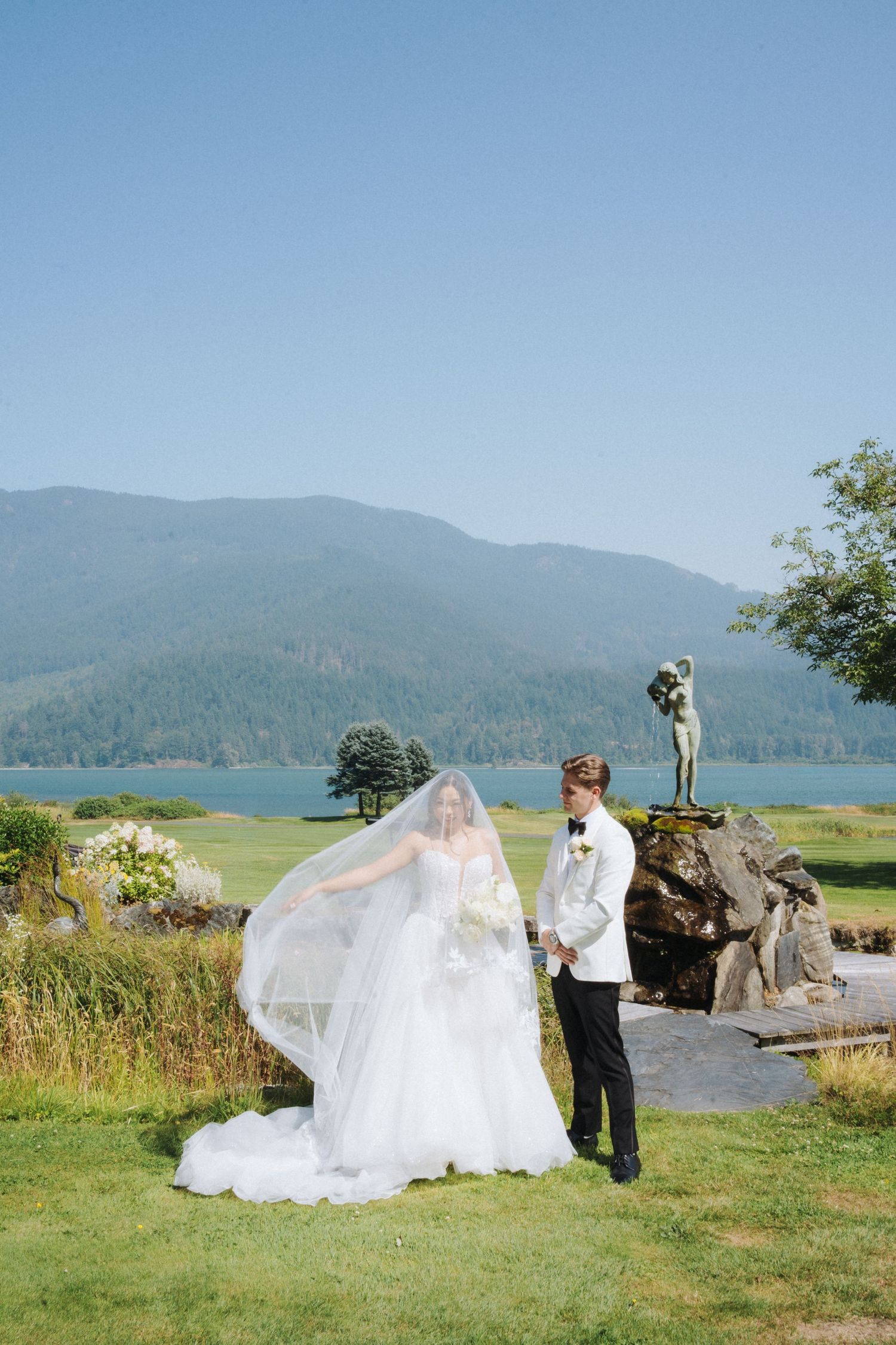Bride and groom pose by a lakeside statue with mountain views, her veil flowing in the breeze on their wedding day.