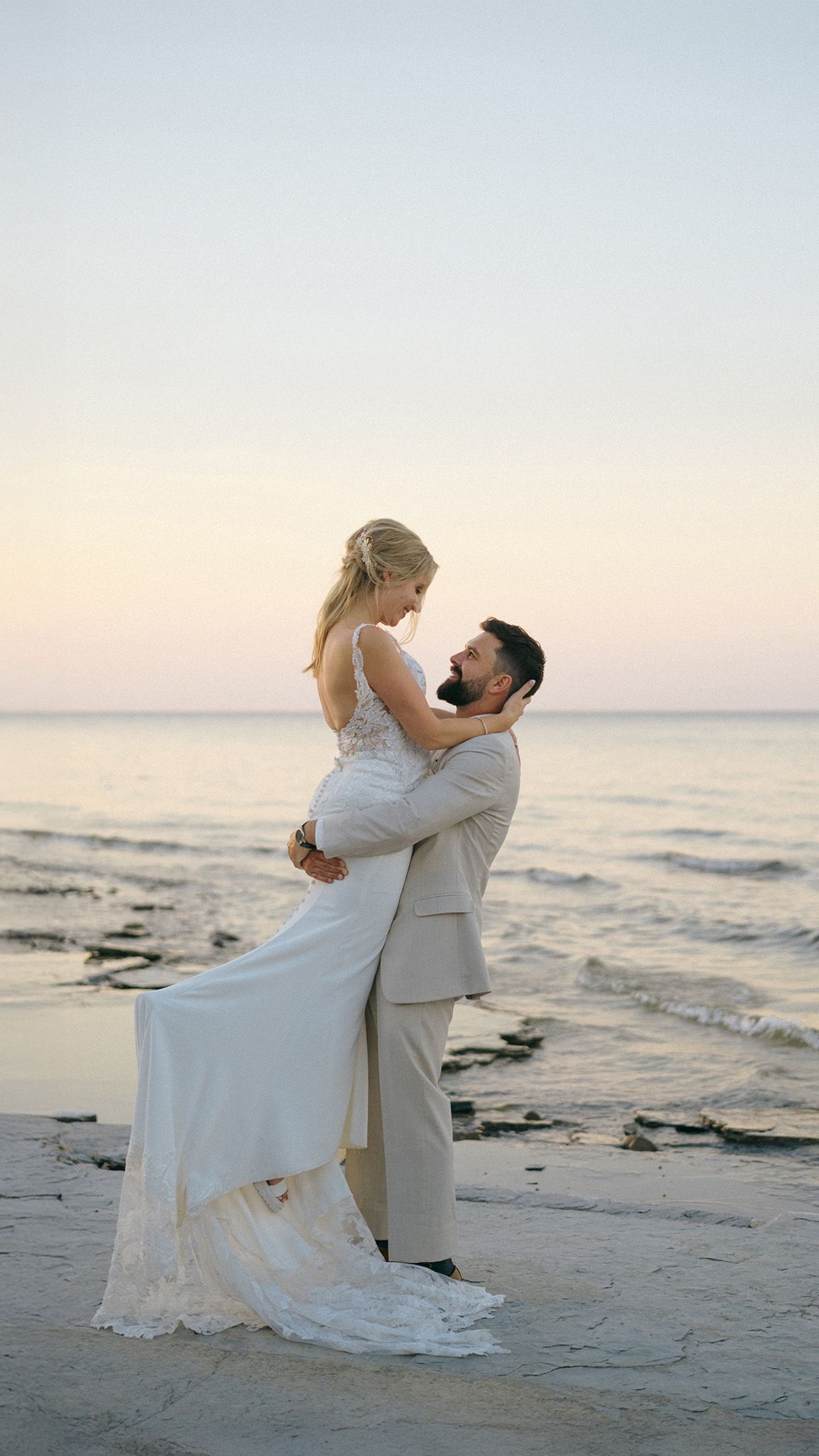 Groom lifts bride in white wedding dress on a serene beach at sunset, sharing a romantic moment by the ocean.