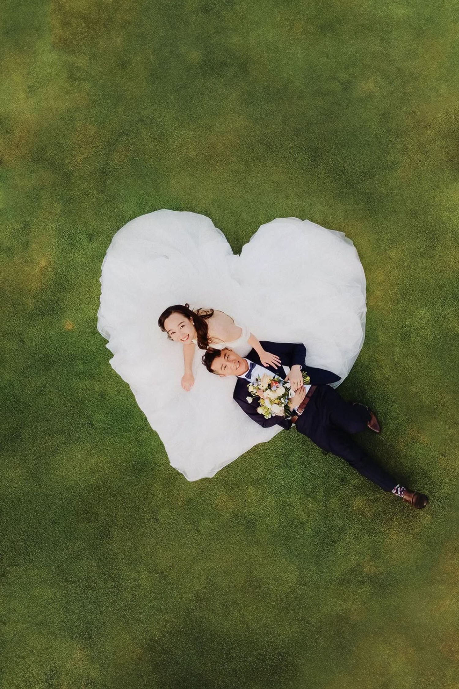 Aerial view of newlyweds lying on green grass, bride's white gown arranged in a heart shape around the couple.