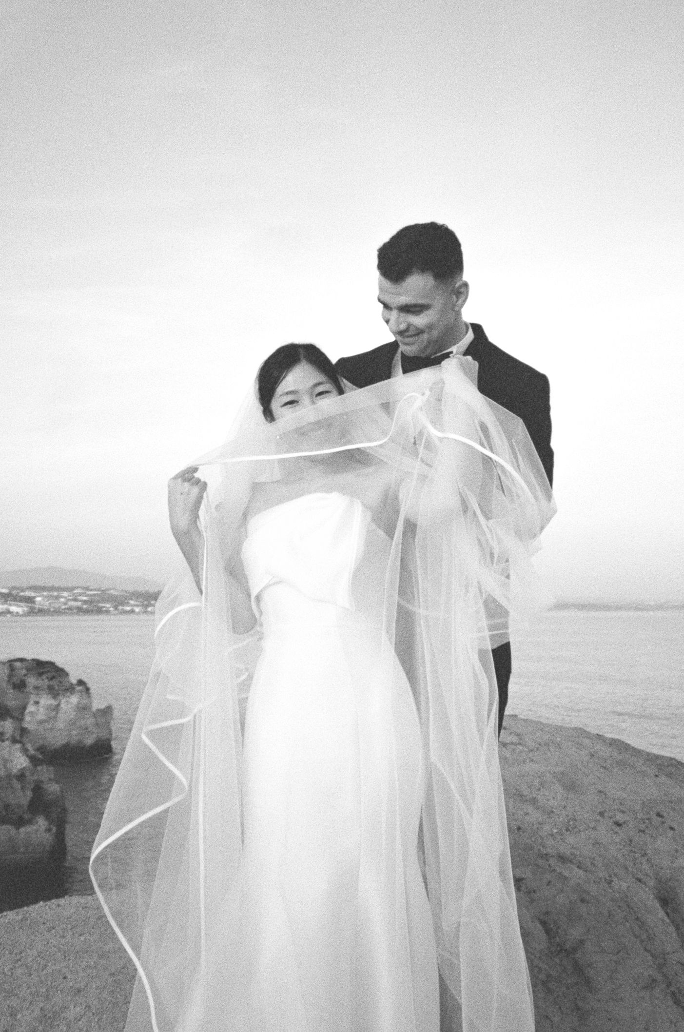 Black and white photo of a bride and groom posing on rocky seaside, her veil flowing in the wind.