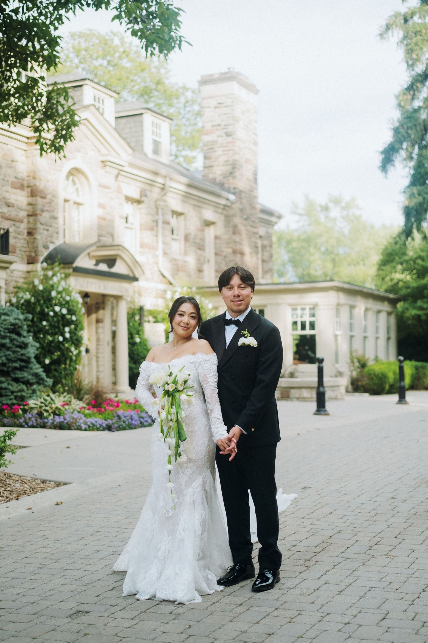 Bride and groom holding hands in front of a stone manor, she in lace gown with bouquet, he in black tuxedo.