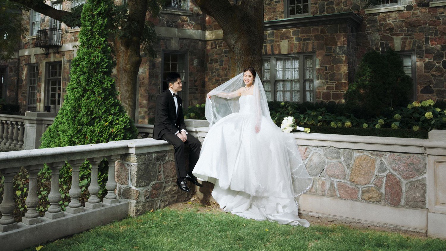 A bride in a white gown and groom in a black suit pose by stone balustrade at an elegant mansion wedding venue.
