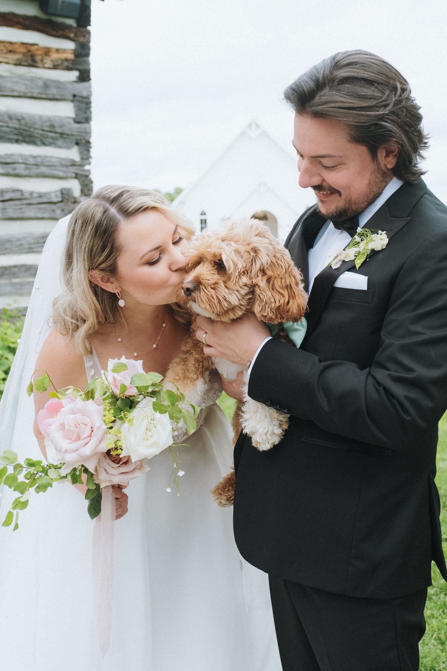 Bride kisses puppy while groom holds dog on wedding day outside white chapel.