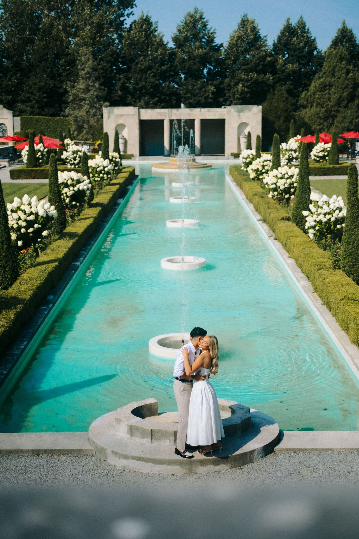 Couple embracing by a turquoise fountain garden with white hydrangeas and manicured hedges at an elegant outdoor venue.
