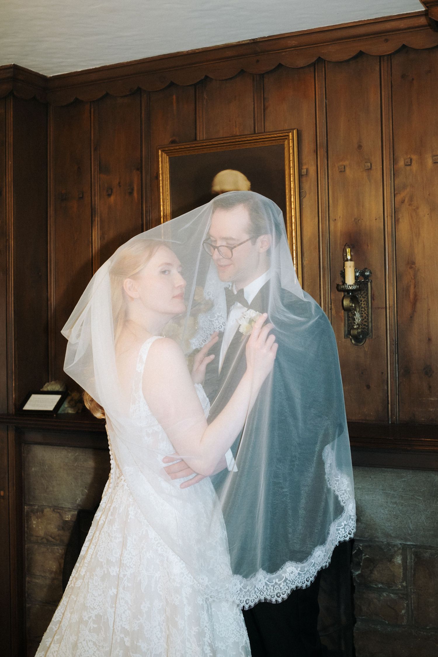 Bride and groom share an intimate moment under a lace wedding veil in an elegant wood-paneled room.