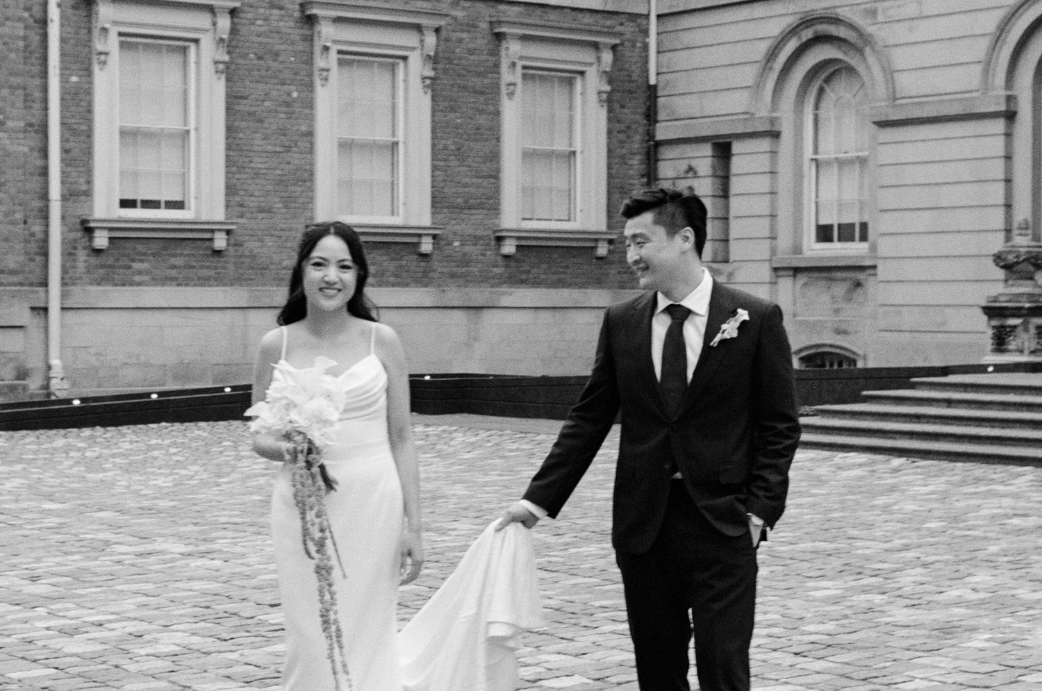 Black and white photo of a bride and groom walking hand in hand outside a historic brick building on their wedding day.