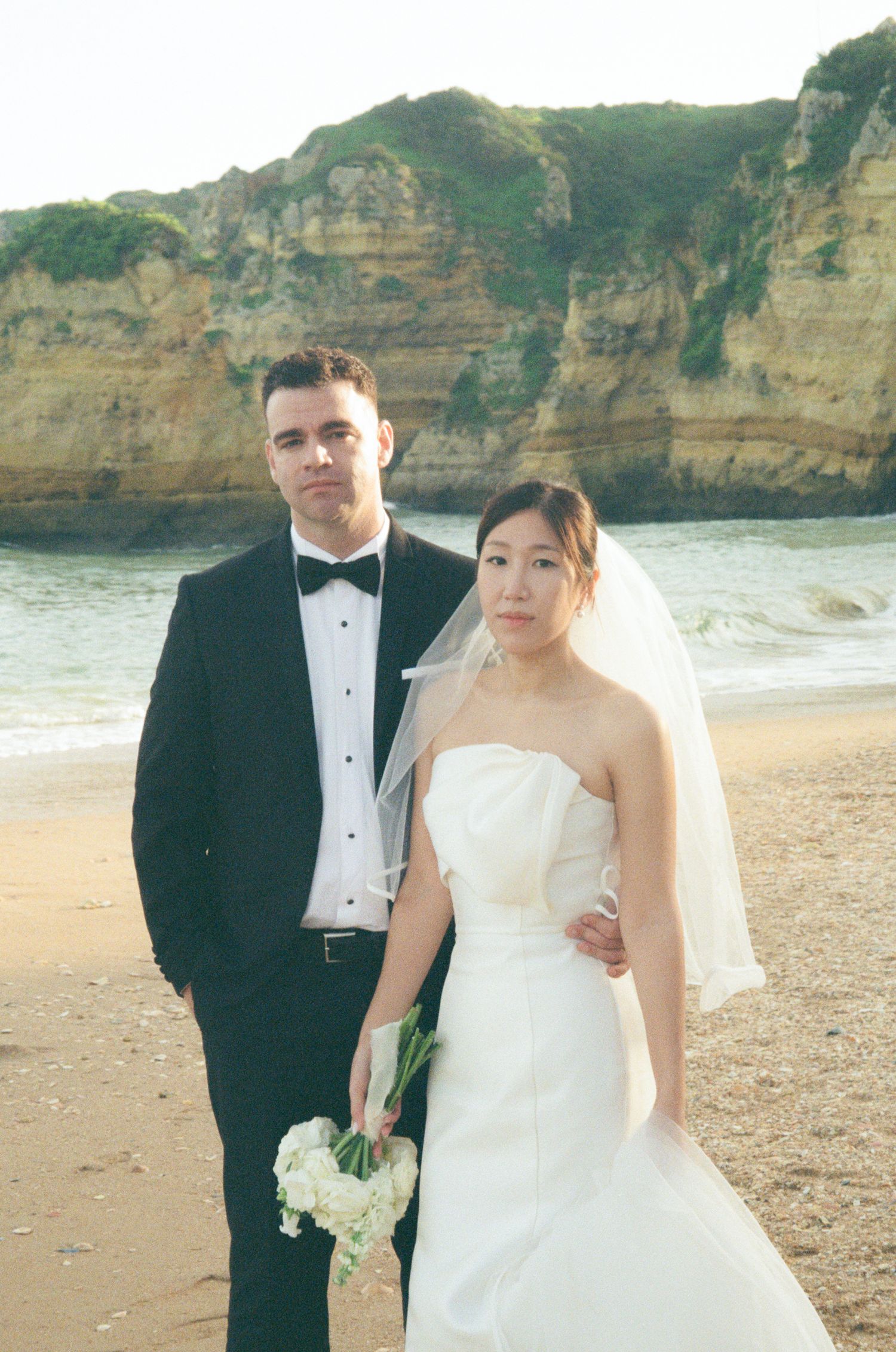 Couple in wedding attire posing on a beach, groom in tuxedo and bride in white gown holding bouquet.