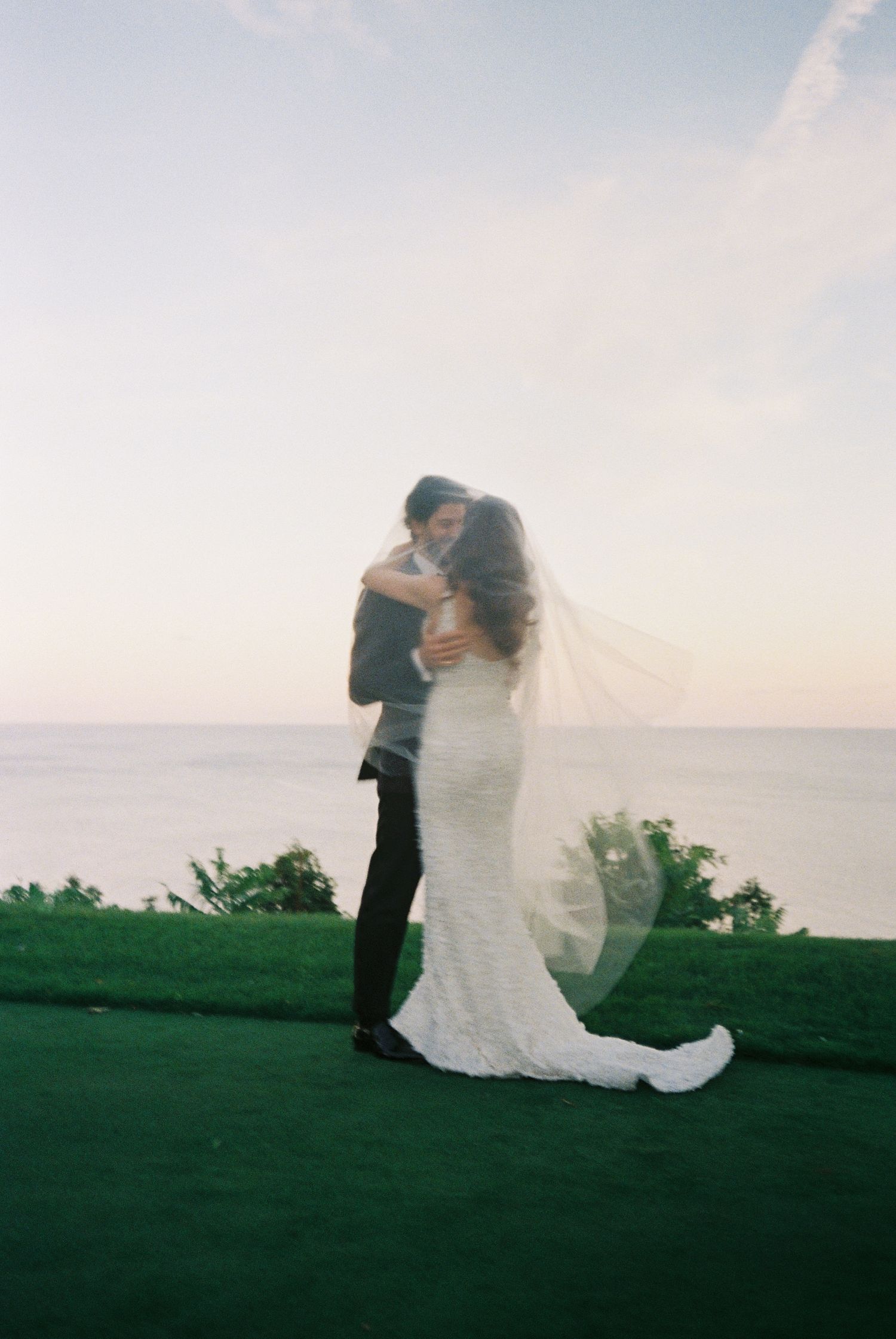 Bride and groom share intimate moment on seaside lawn at dusk with flowing veil.