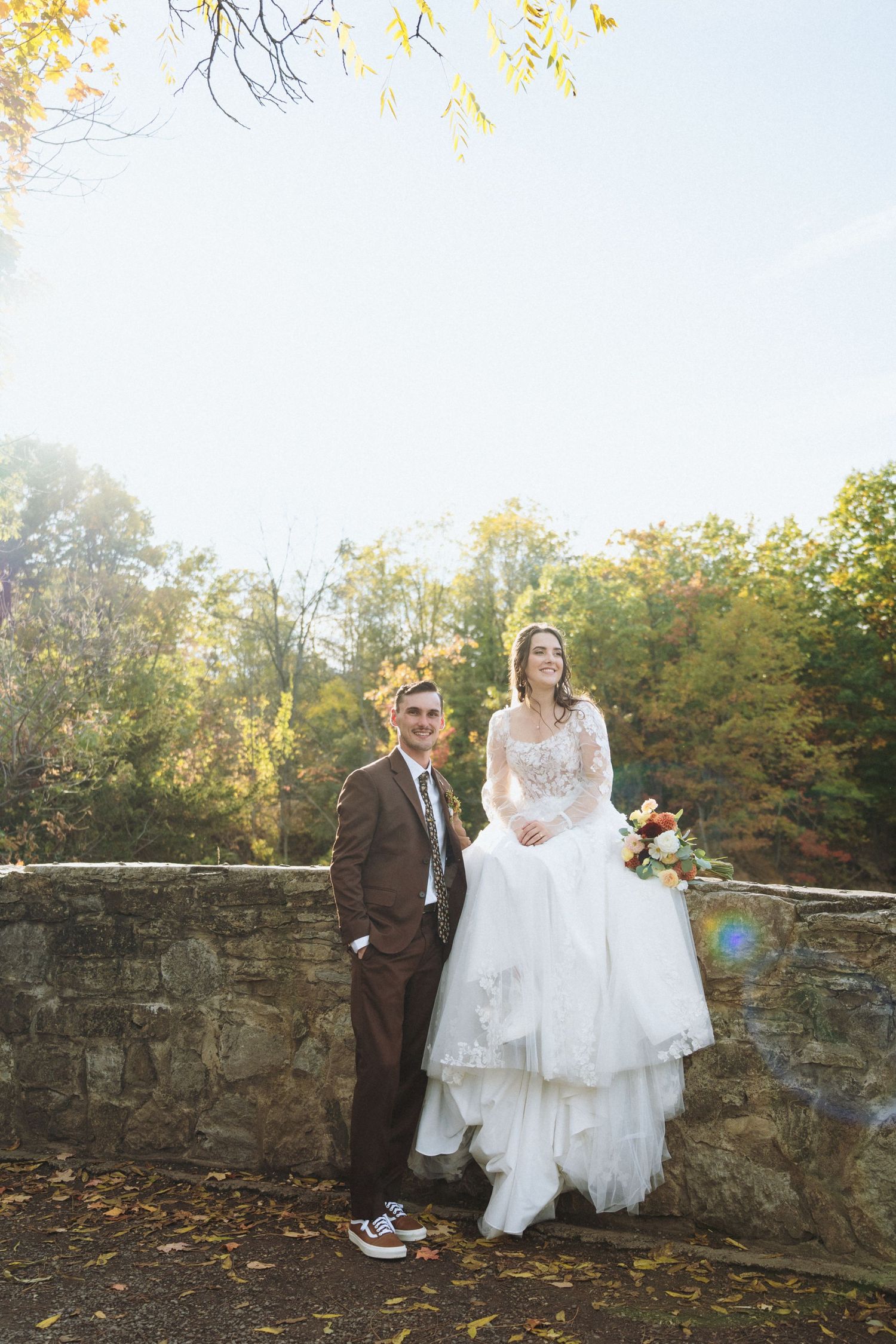 Bride in lace gown sits on stone wall beside groom in brown suit amid autumn foliage.
