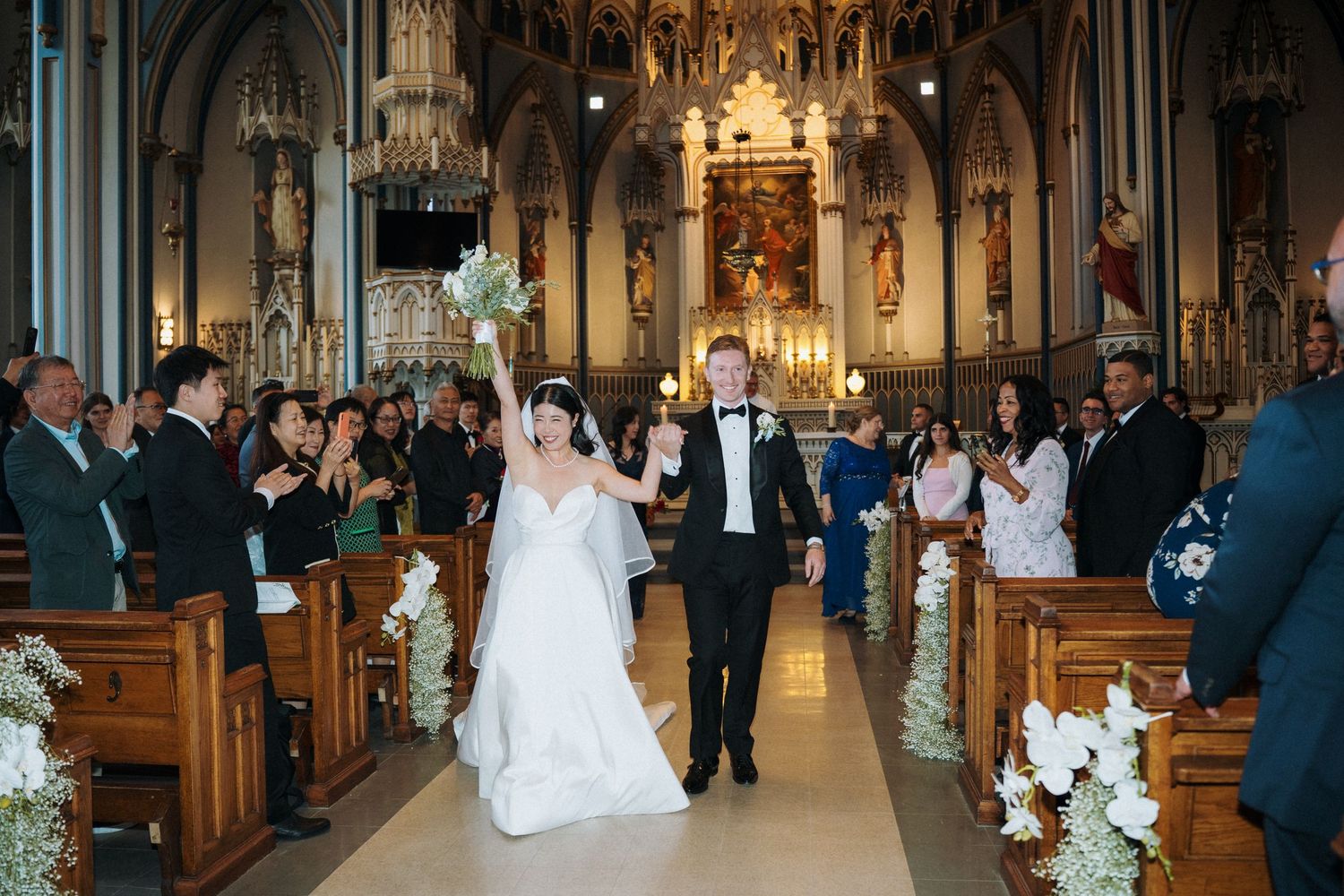 Bride and groom walk down church aisle after wedding ceremony, guests applauding in ornate Catholic cathedral.