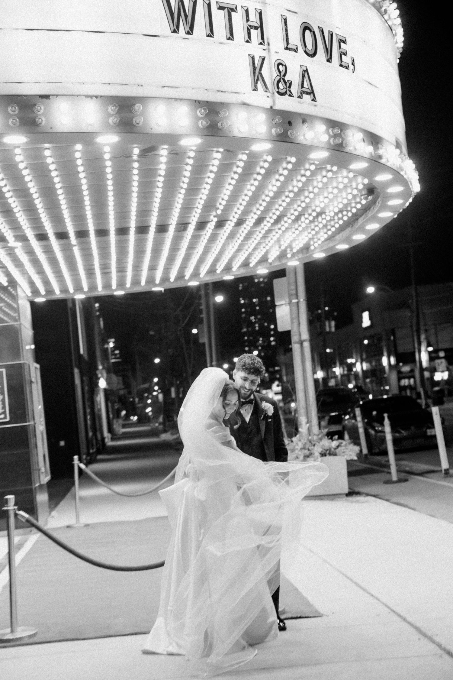 Black and white photo of bride and groom kissing under a lit marquee sign reading With Love K&A at night.