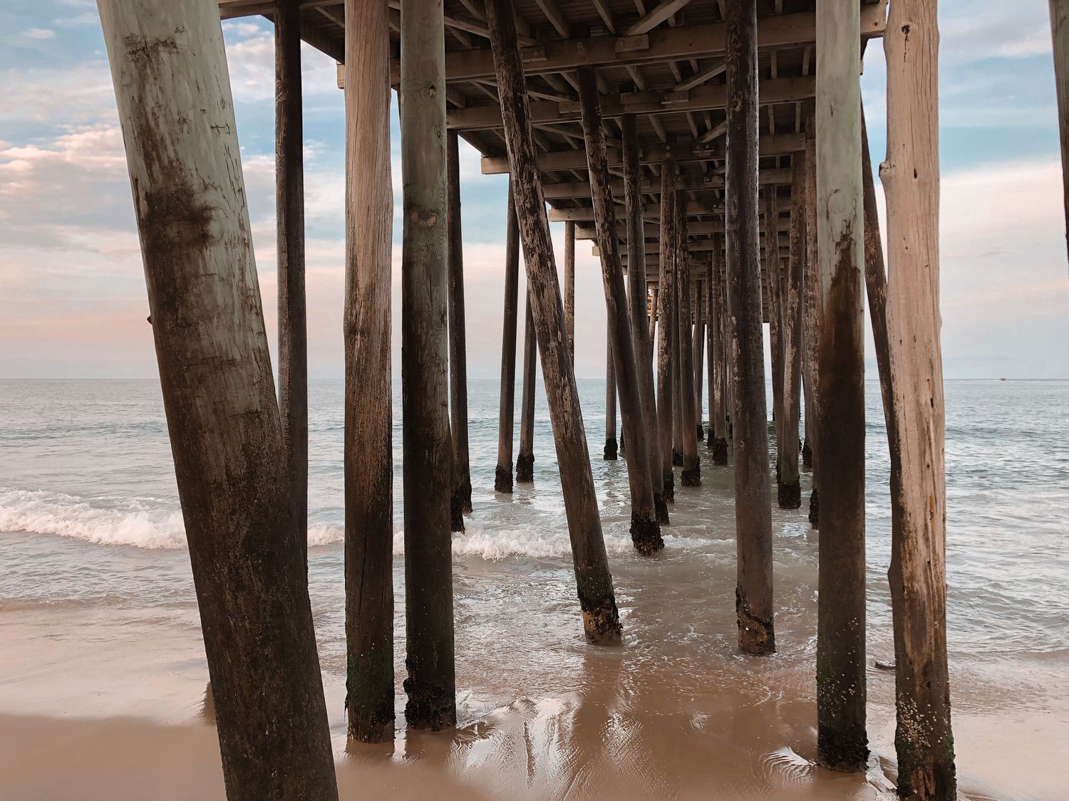 Wooden pier pilings create geometric patterns while stretching into the ocean at sunset on a sandy beach.