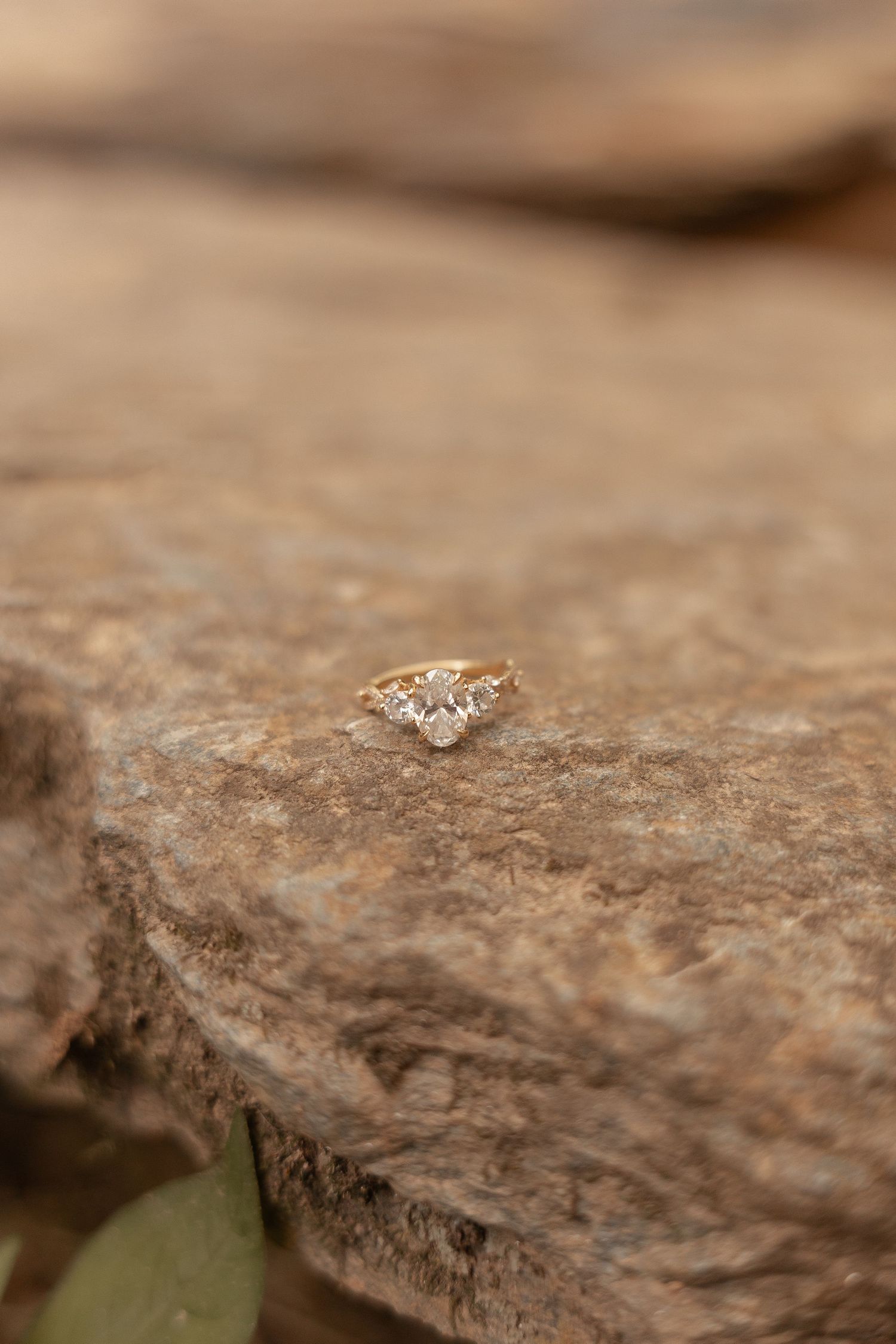 Gold diamond engagement ring with three stones resting on a textured rock surface outdoors.