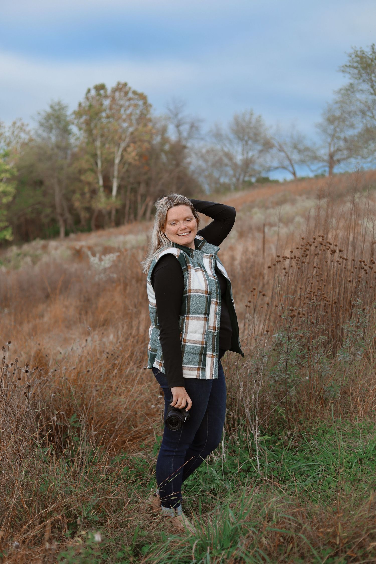 A person wearing a plaid shirt and black vest explores a field of tall brown grass on an autumn day.